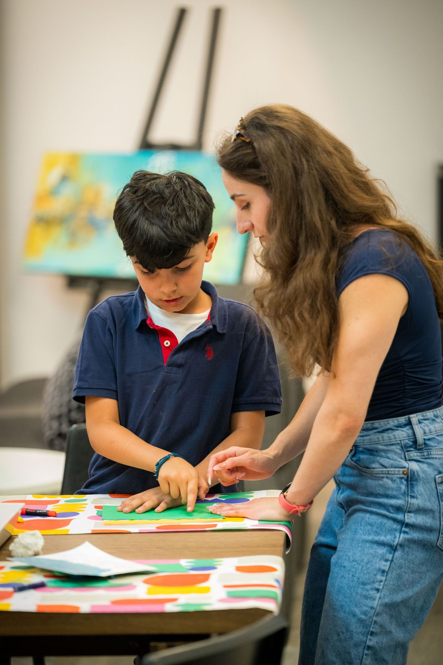 Teacher helps young boy arrange colorful paper cutouts on green paper during art class activity.