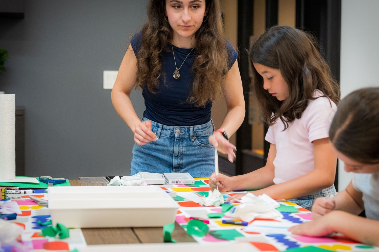 A female teacher supervises two young girls creating colorful paper art at a table.