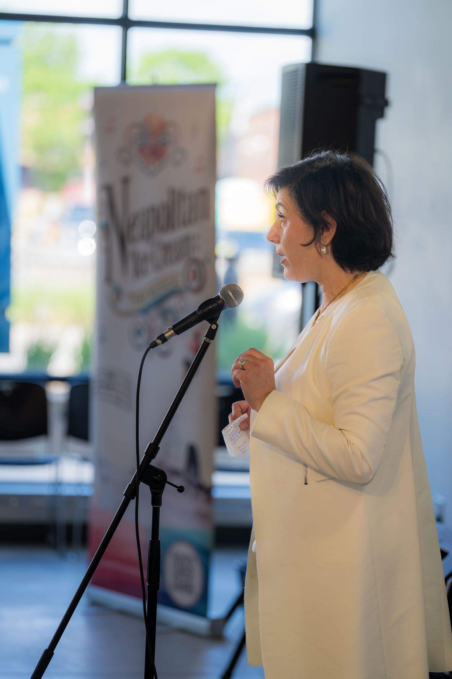 Woman in white outfit speaking at microphone stand at Neapolitan-themed event.