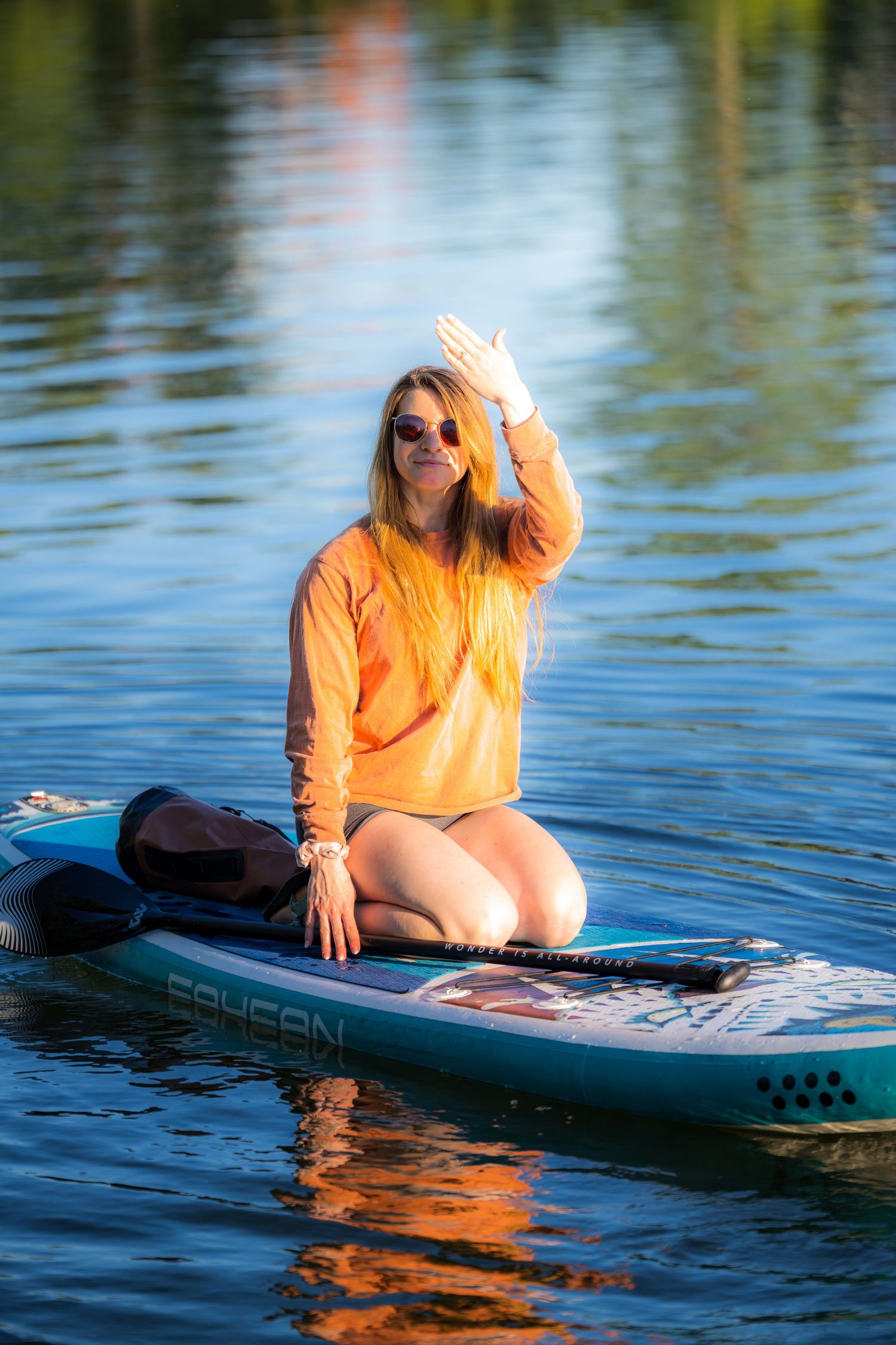 Woman in orange hoodie sitting on a paddleboard on calm water, shielding her eyes from the sun.