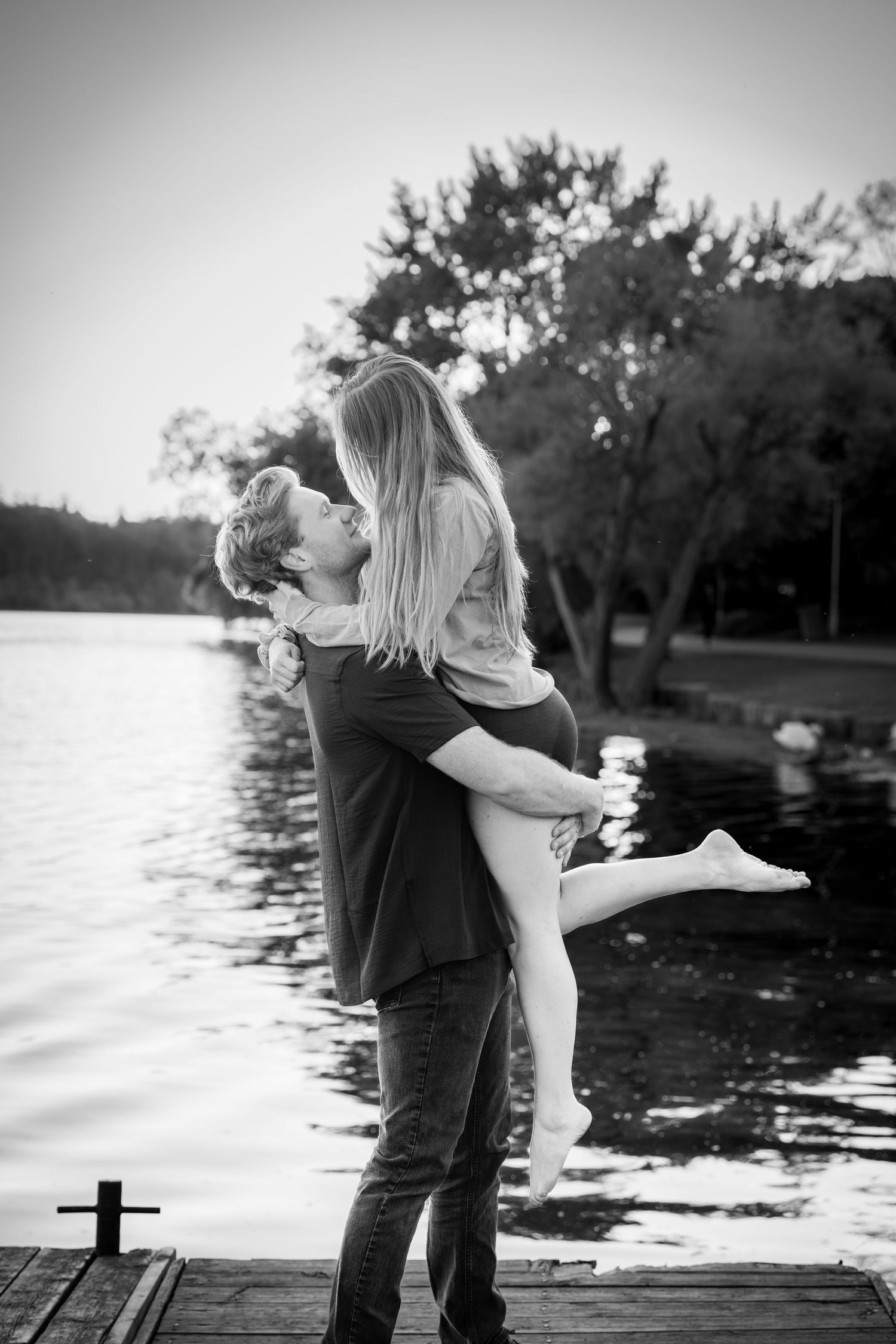 Black and white photo of man lifting woman on a wooden dock by a lake in a romantic pose.