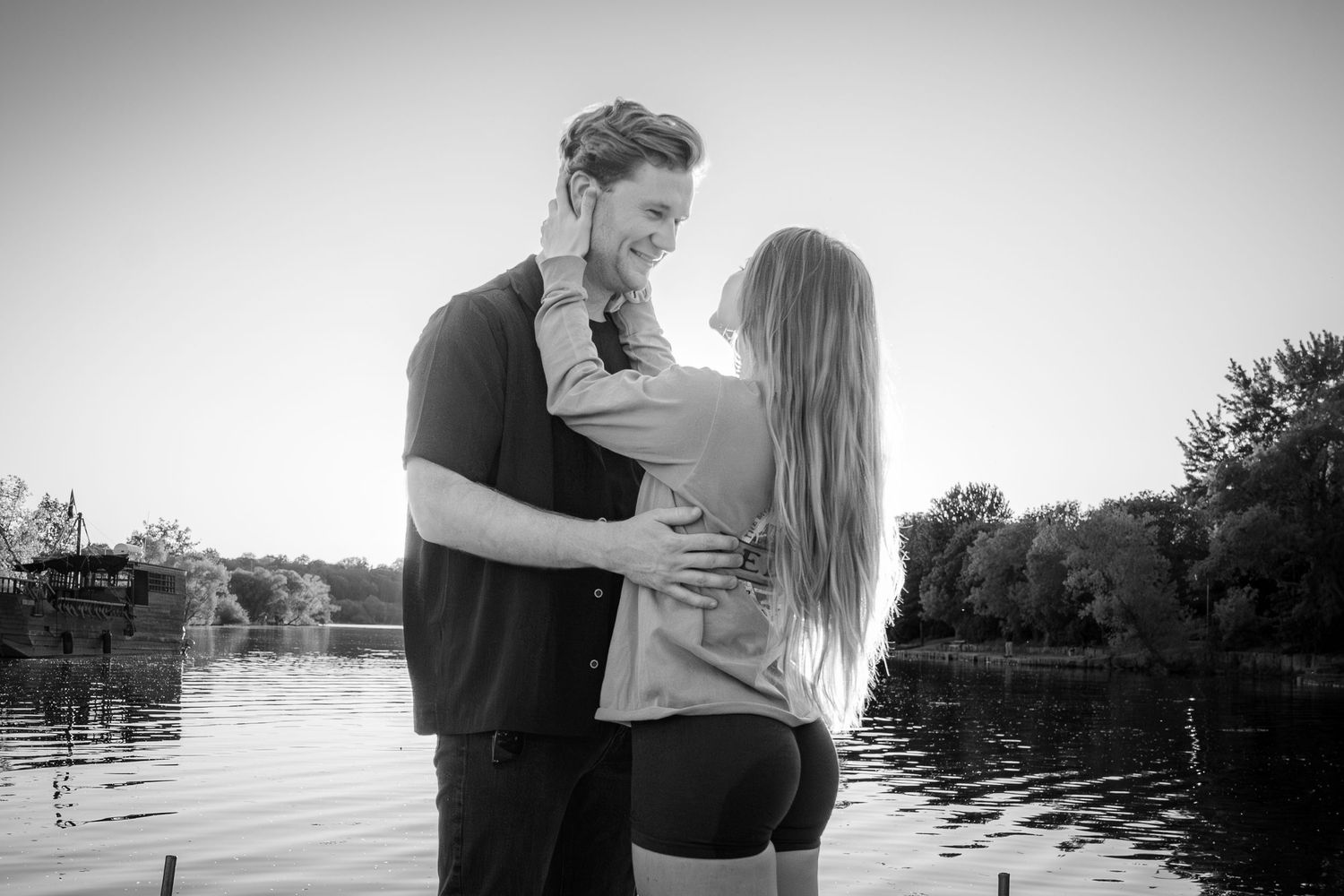 Black and white photo of romantic couple embracing by a peaceful lake at sunset.