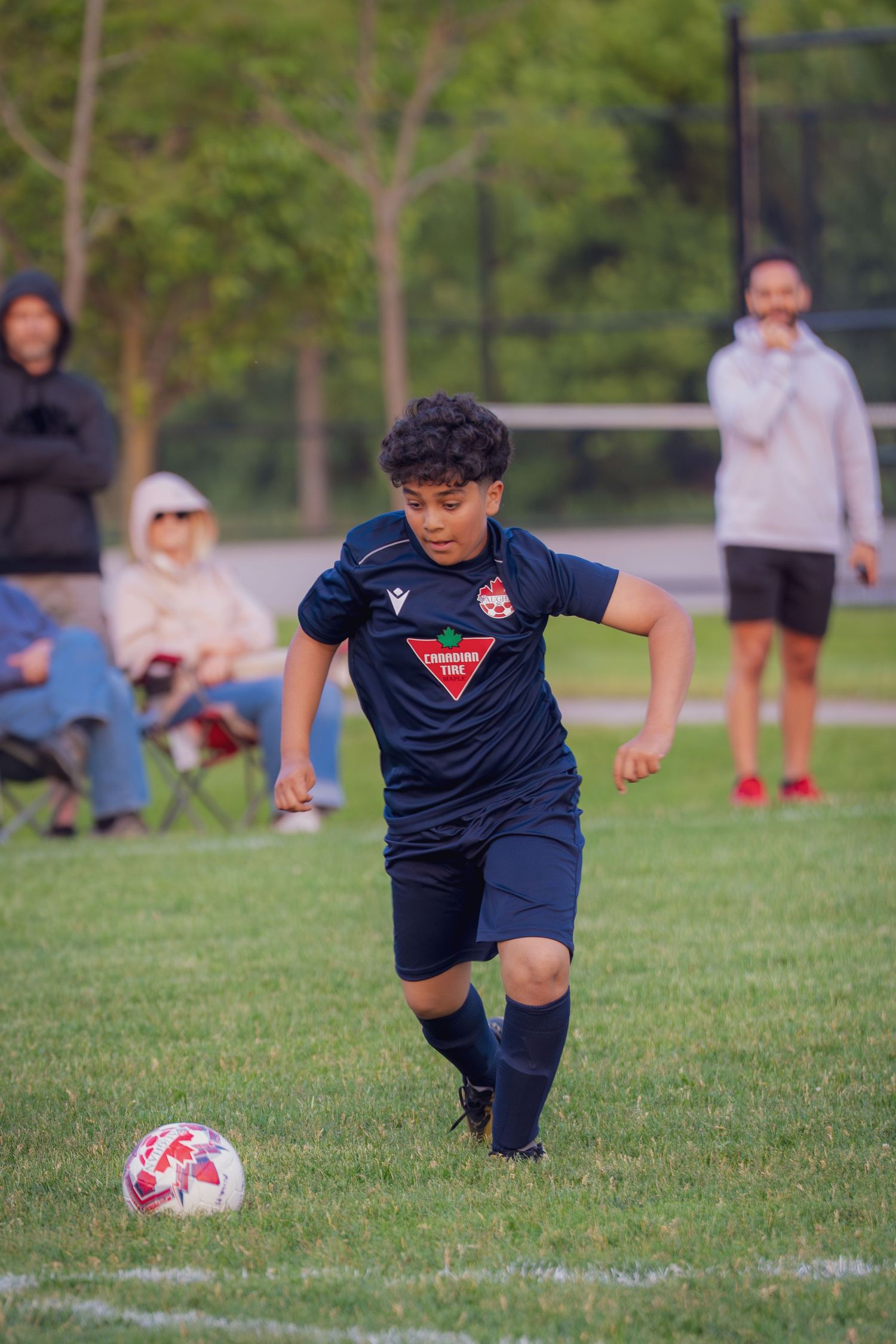 Soccer player in dark uniform practices kicking drills on grass field with spectators in background.