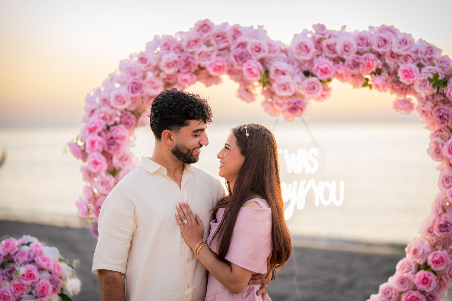 Heart-shaped pink flower arch on beach with ocean backdrop during golden hour.