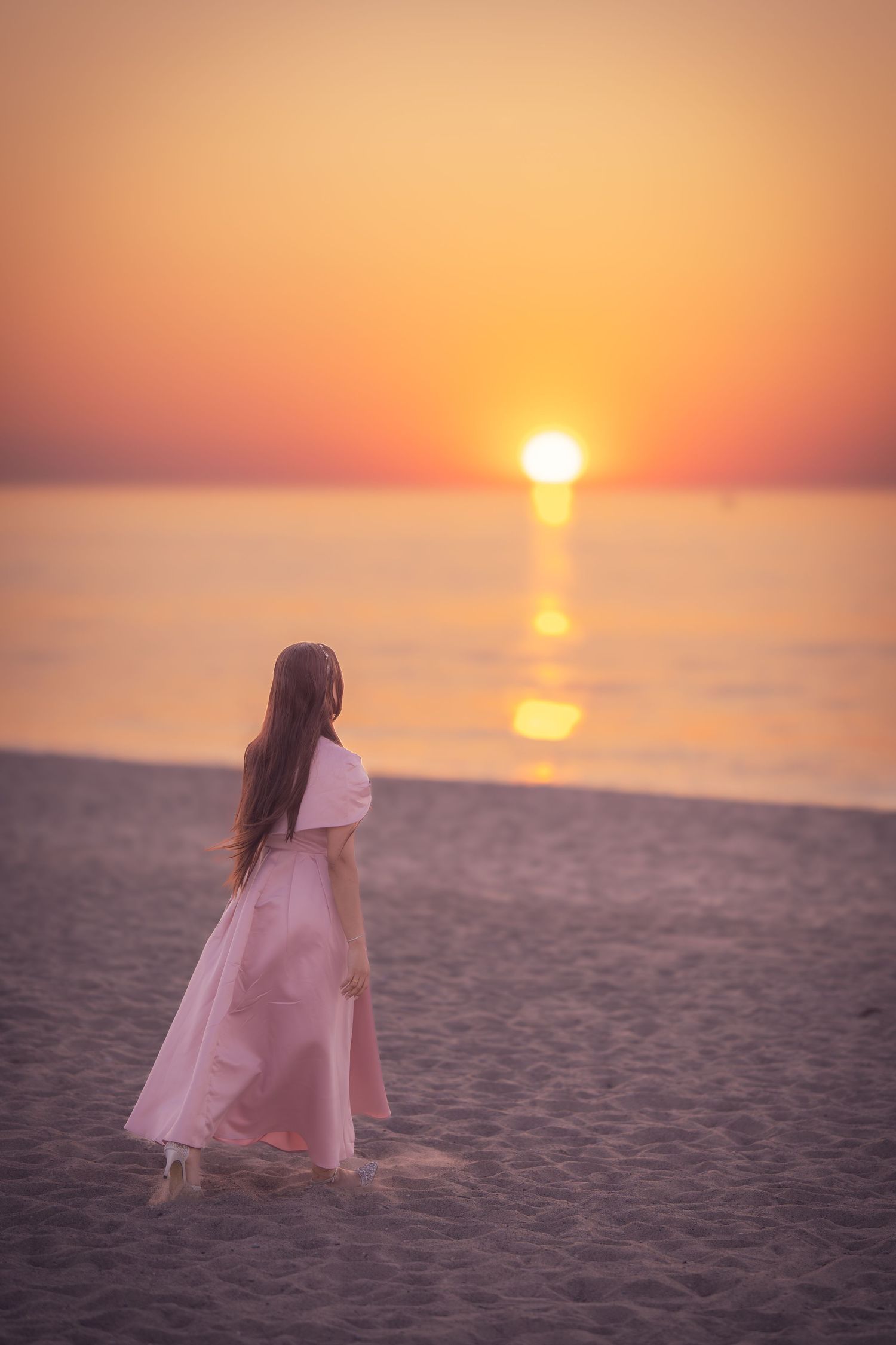 A figure in a flowing pink dress walks on a sandy beach at sunset with golden light reflecting on the ocean.