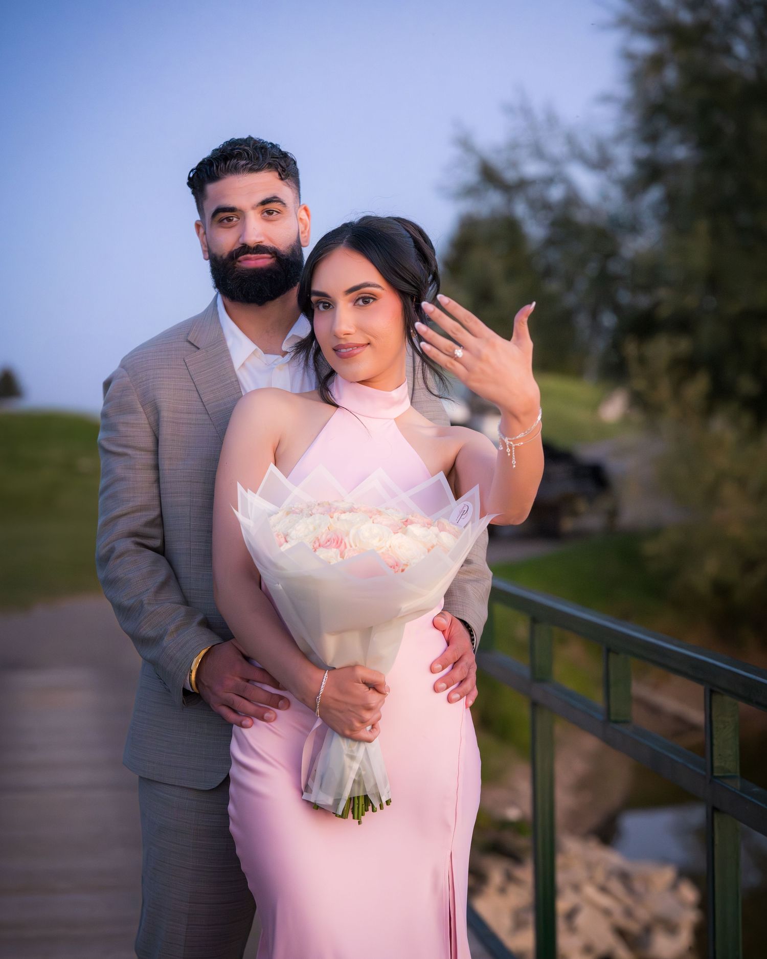 A couple poses romantically on a path by a lake at sunset, with the person in a pink dress holding a white bouquet.