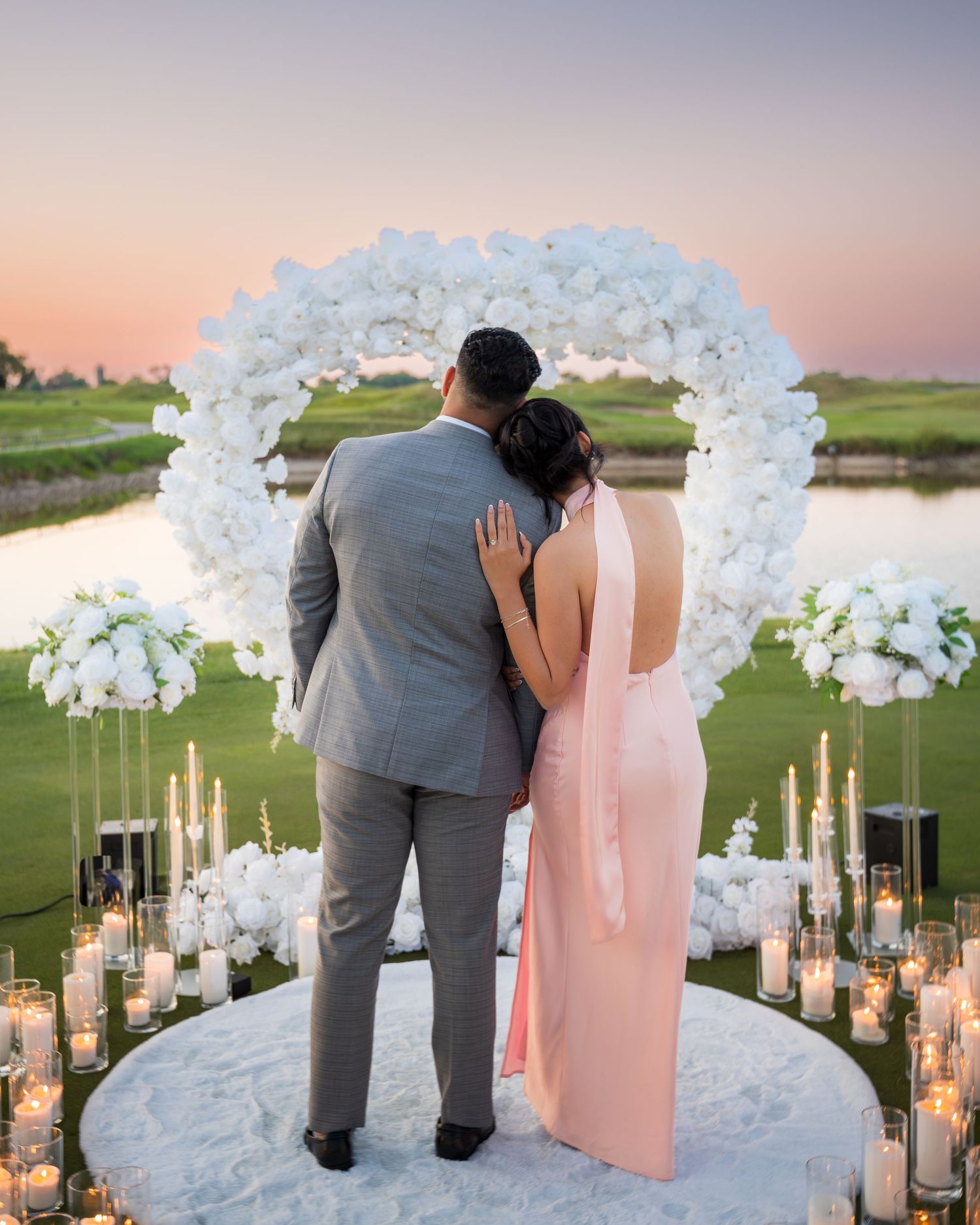 Wedding couple stands beneath white floral arch with glowing candles at sunset.