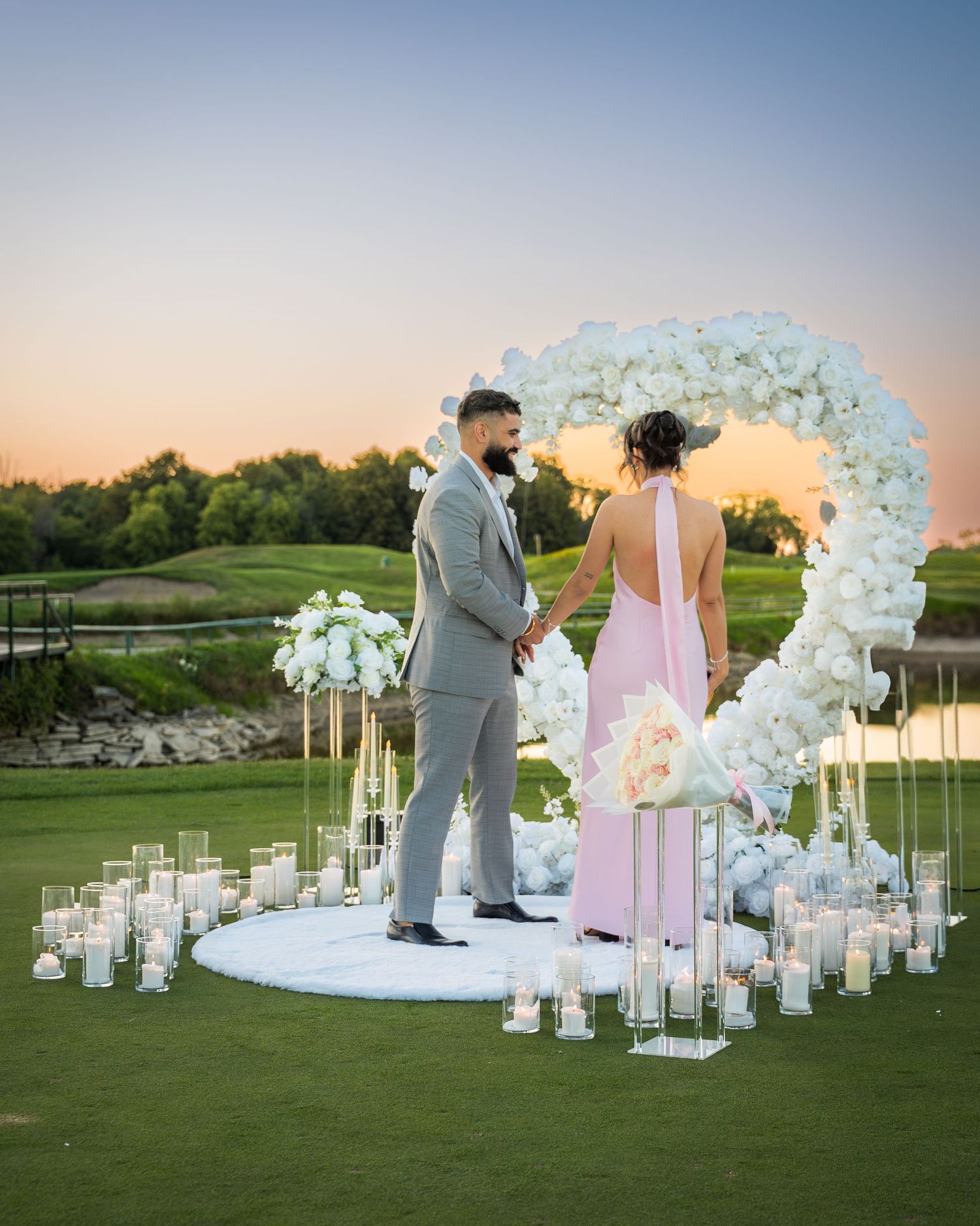 Elegant outdoor proposal scene with white floral arches and lanterns on a manicured lawn at dusk.