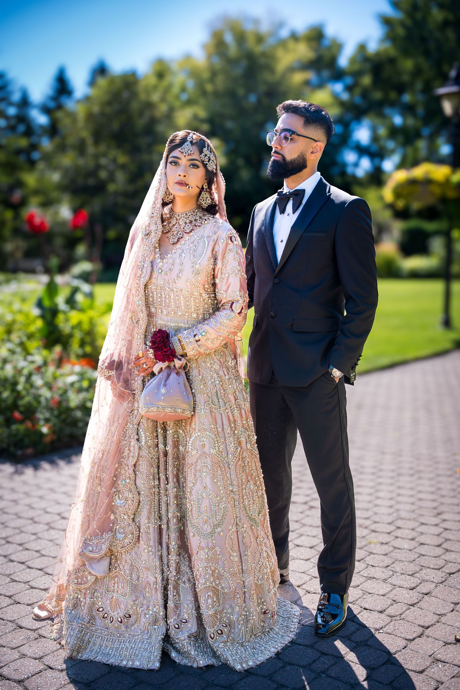 A couple in formal attire poses on a brick pathway surrounded by garden scenery.