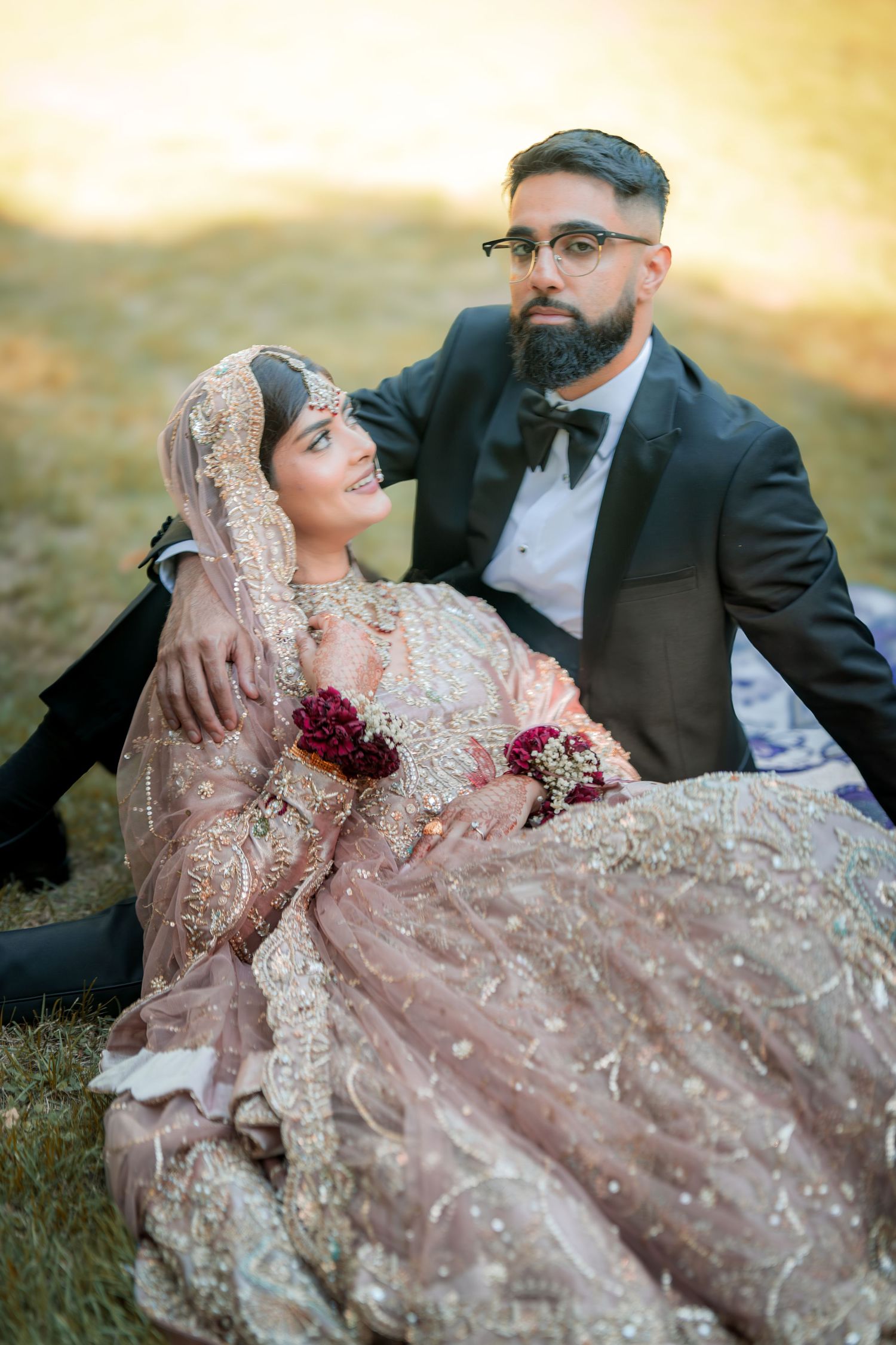 Intimate outdoor wedding photo of couple in formal South Asian wedding attire sitting on grass.