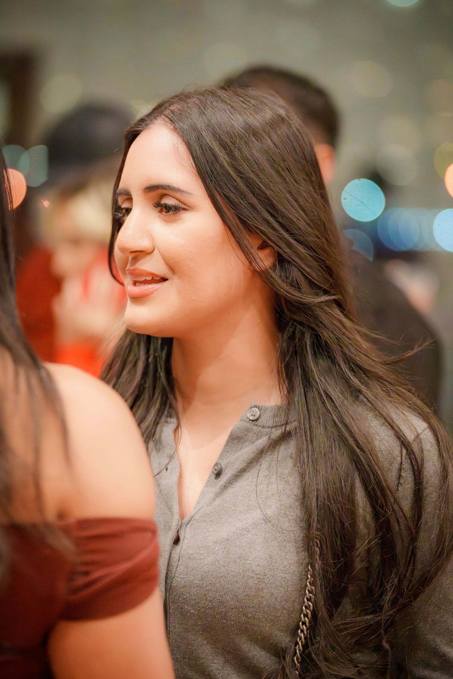 A person with long dark hair wearing a grey top smiles warmly in a dimly lit room.
