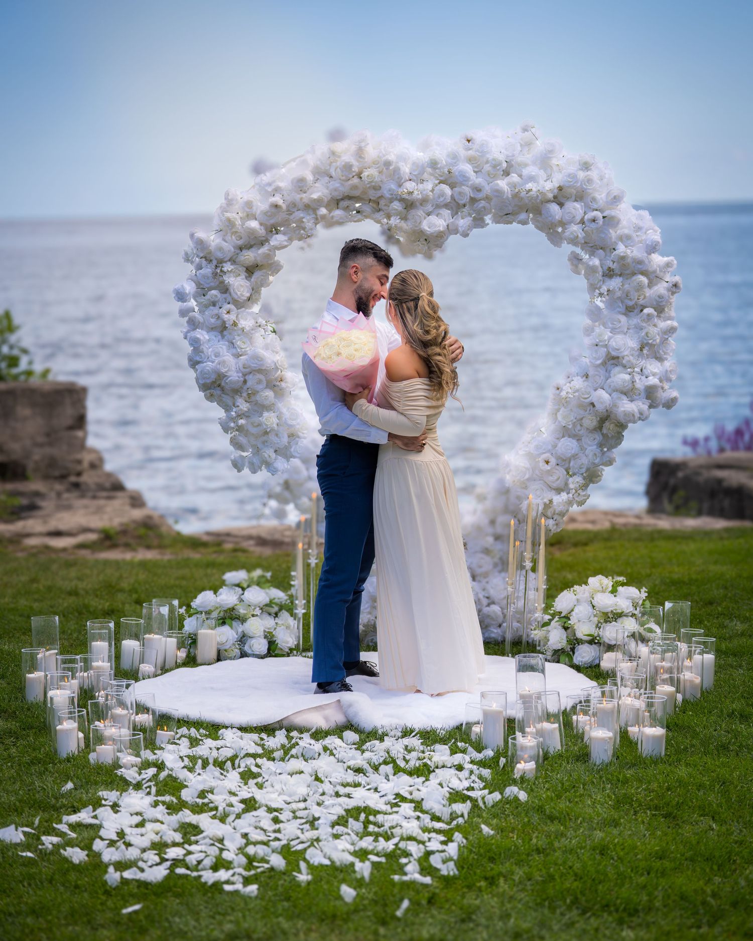 Romantic oceanside wedding ceremony setup with white floral arch and scattered candles on grass.