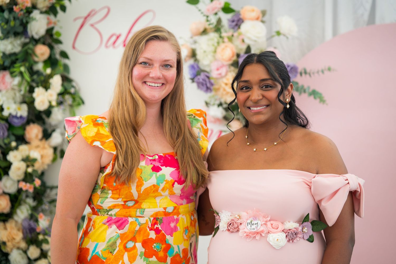 Guests pose together at a baby shower with floral decorations and a pink backdrop.