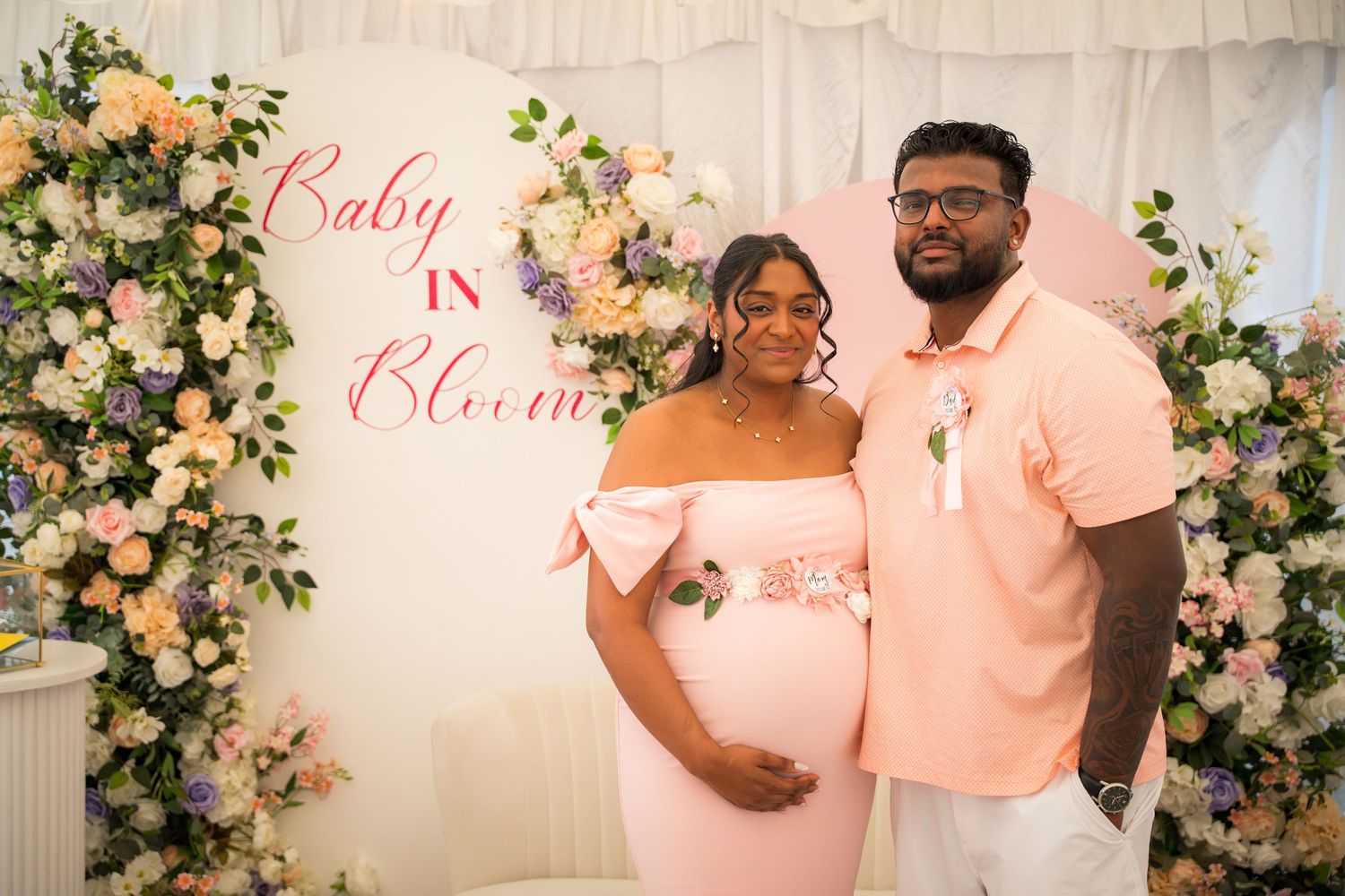 Couple posing at baby shower event with pink and white floral decorations.
