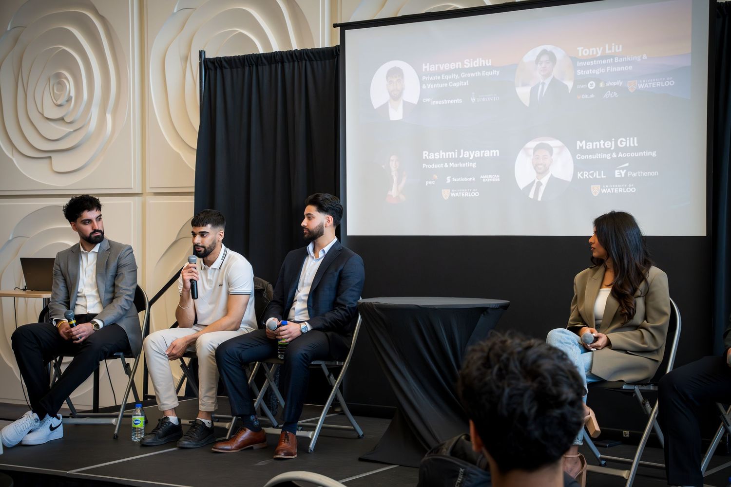 Business panel discussion with presentation screen displaying speaker information in background.
