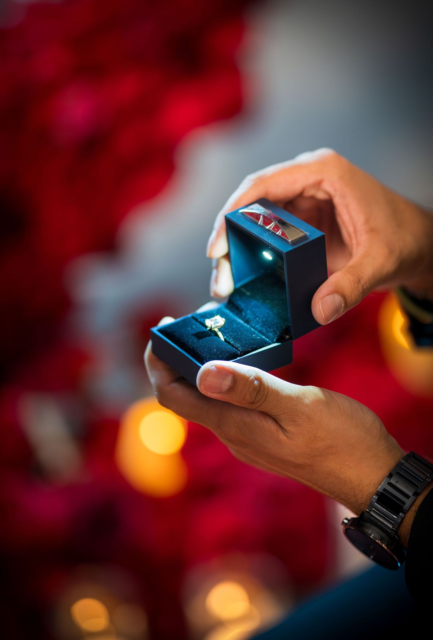 Hands holding a small blue ring box against a backdrop of red roses and candlelight.