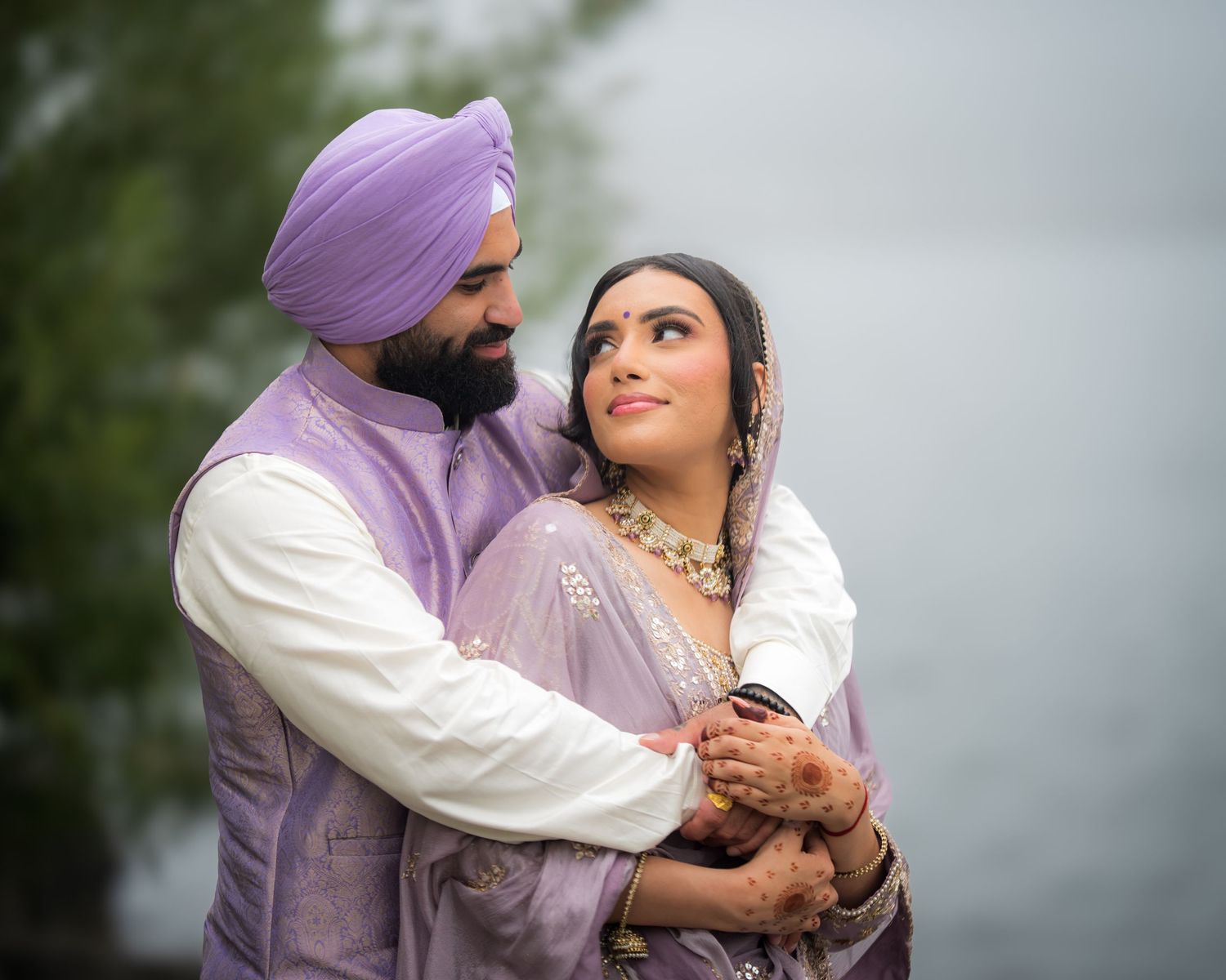 Romantic couple in traditional purple and white attire embrace against cloudy sky.