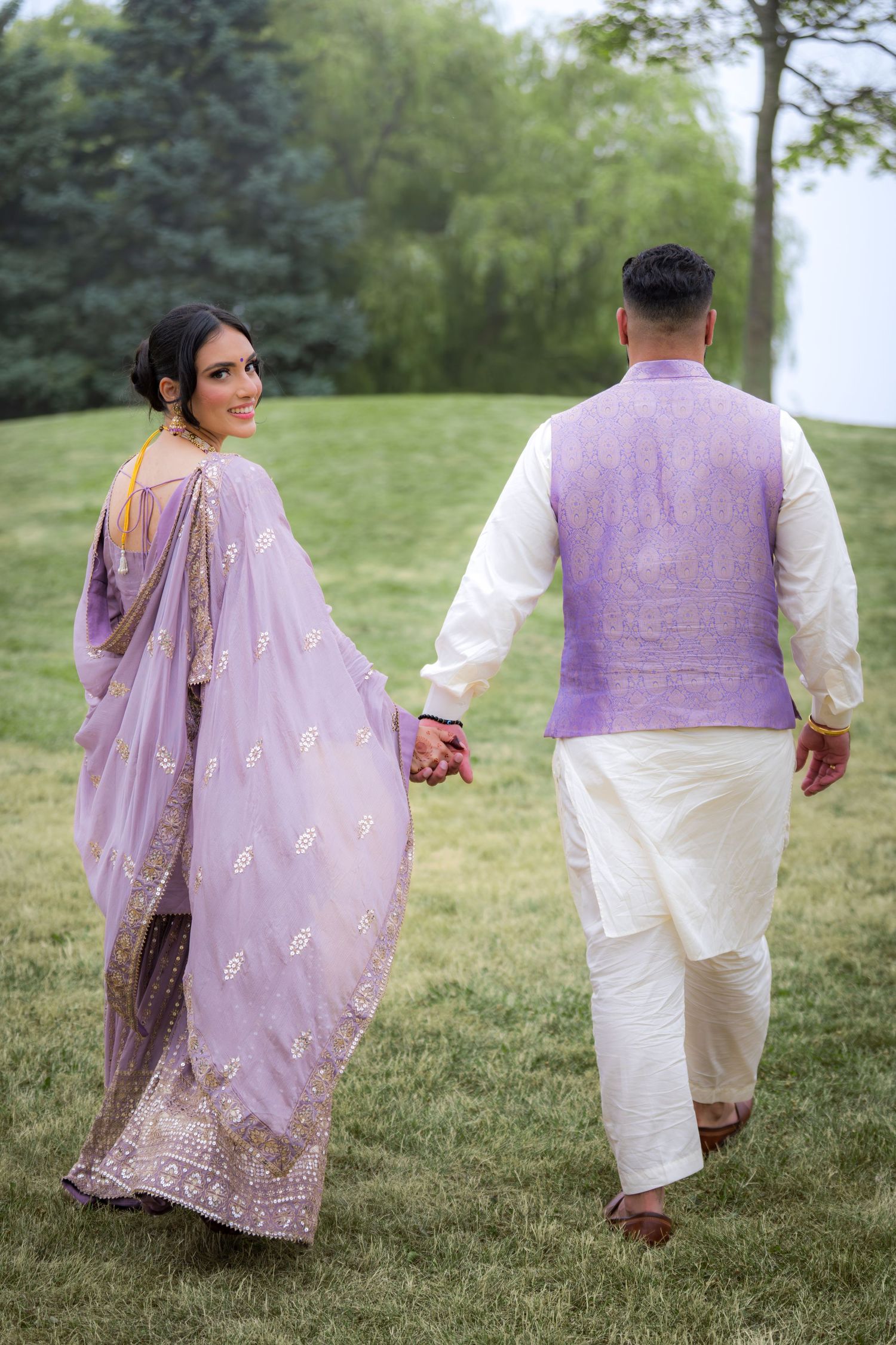 Couple in matching purple and white traditional outfits walking hand in hand through a grassy park.