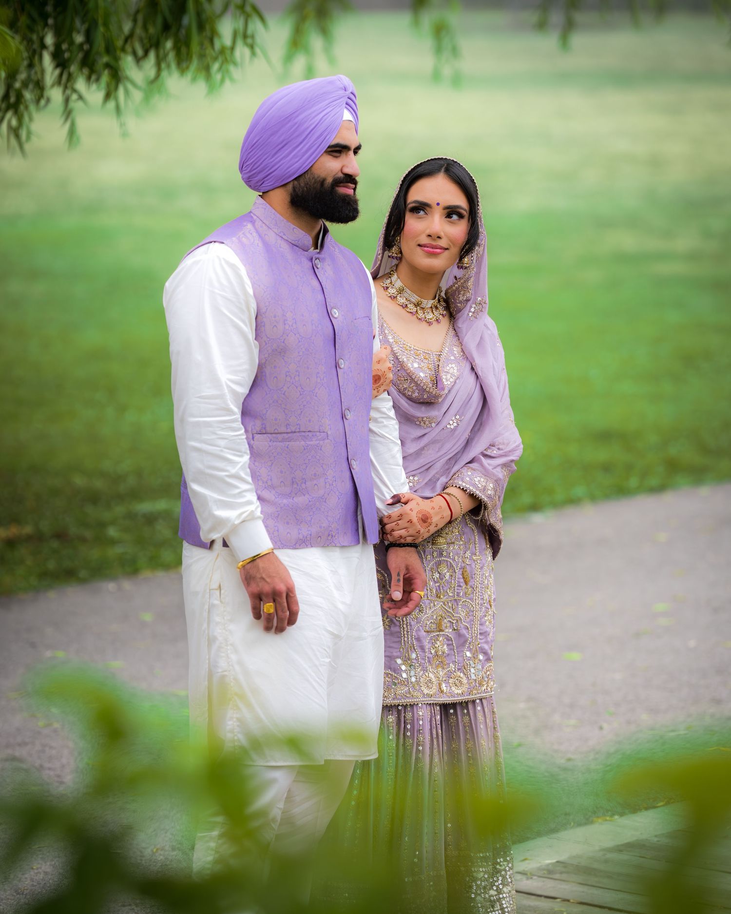 A couple in matching lavender outfits standing on a garden path framed by willow branches.