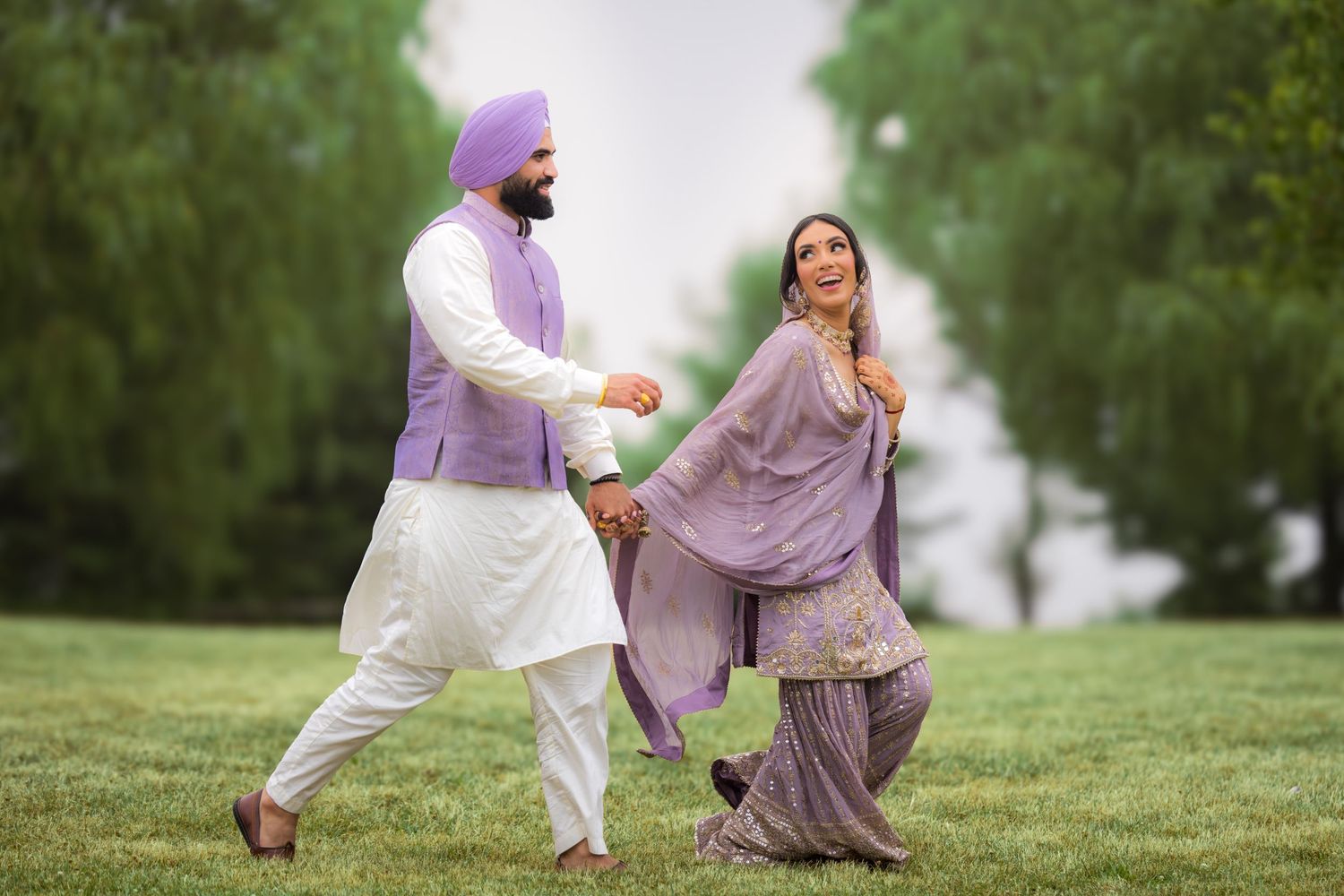 Couple in traditional dress chases each other playfully on a green hillside during photoshoot.