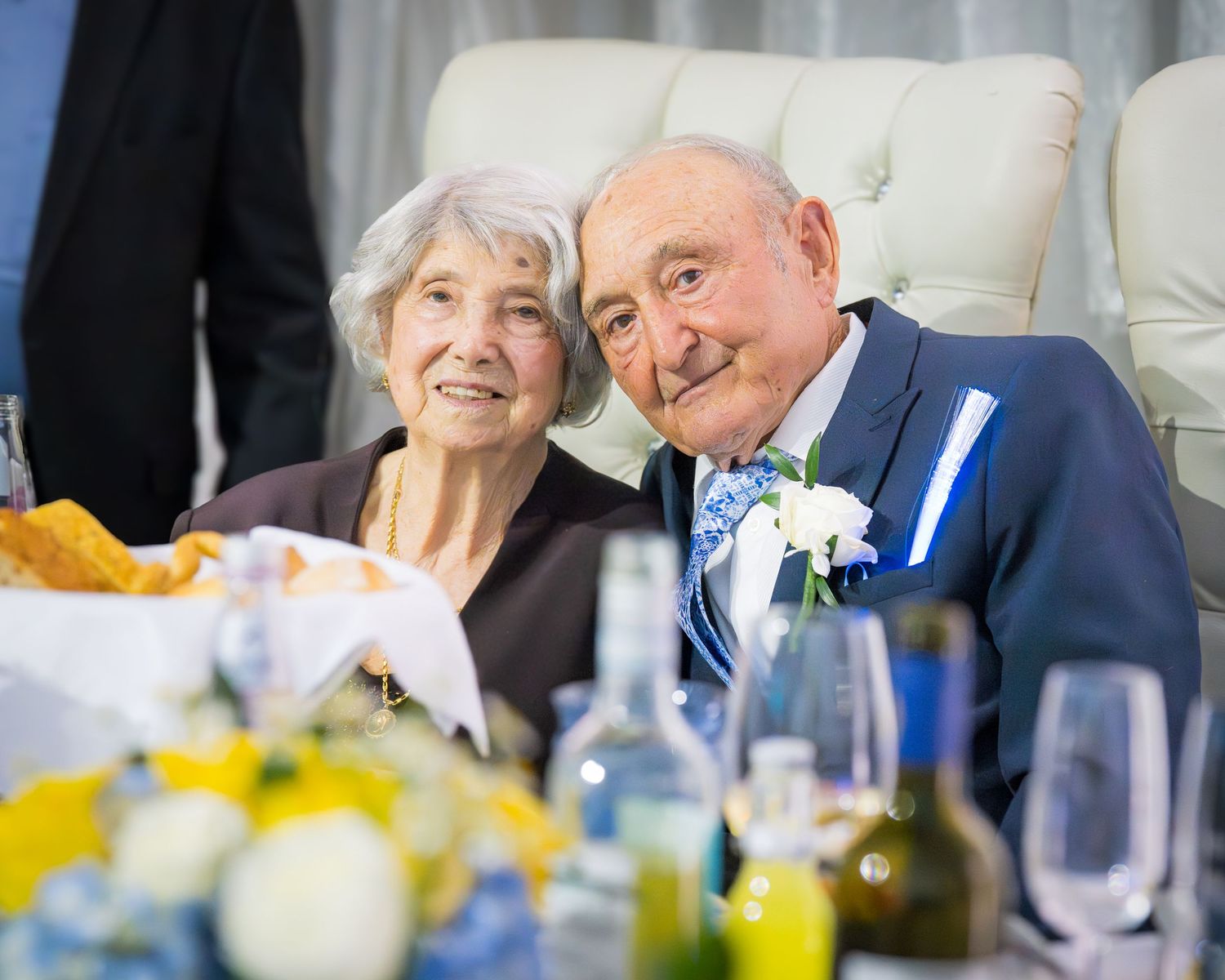 Happy elderly couple posing together at wedding anniversary reception with floral arrangements.