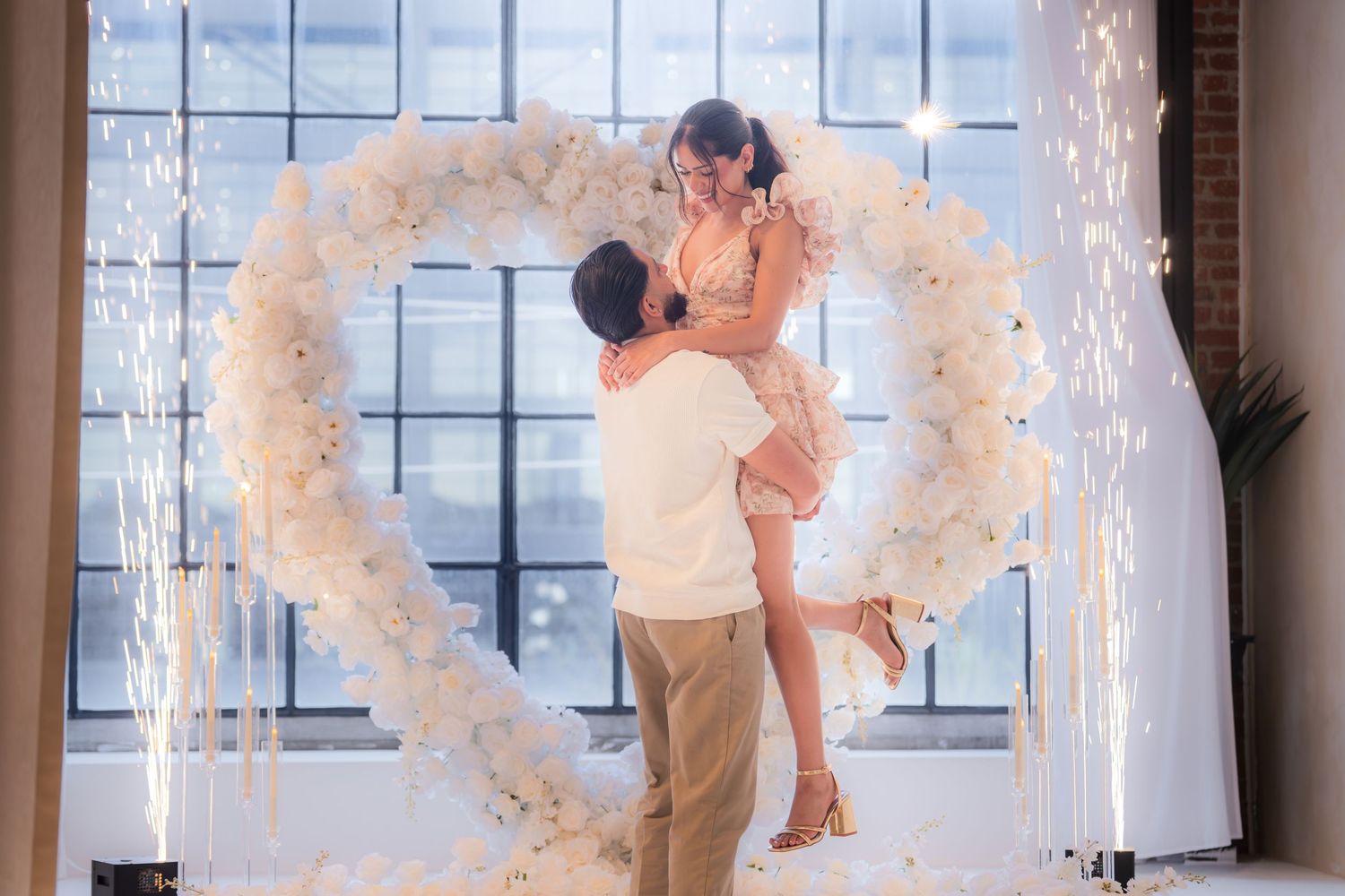 Man lifting woman joyfully in front of heart-shaped white floral arch with sparkling fireworks.