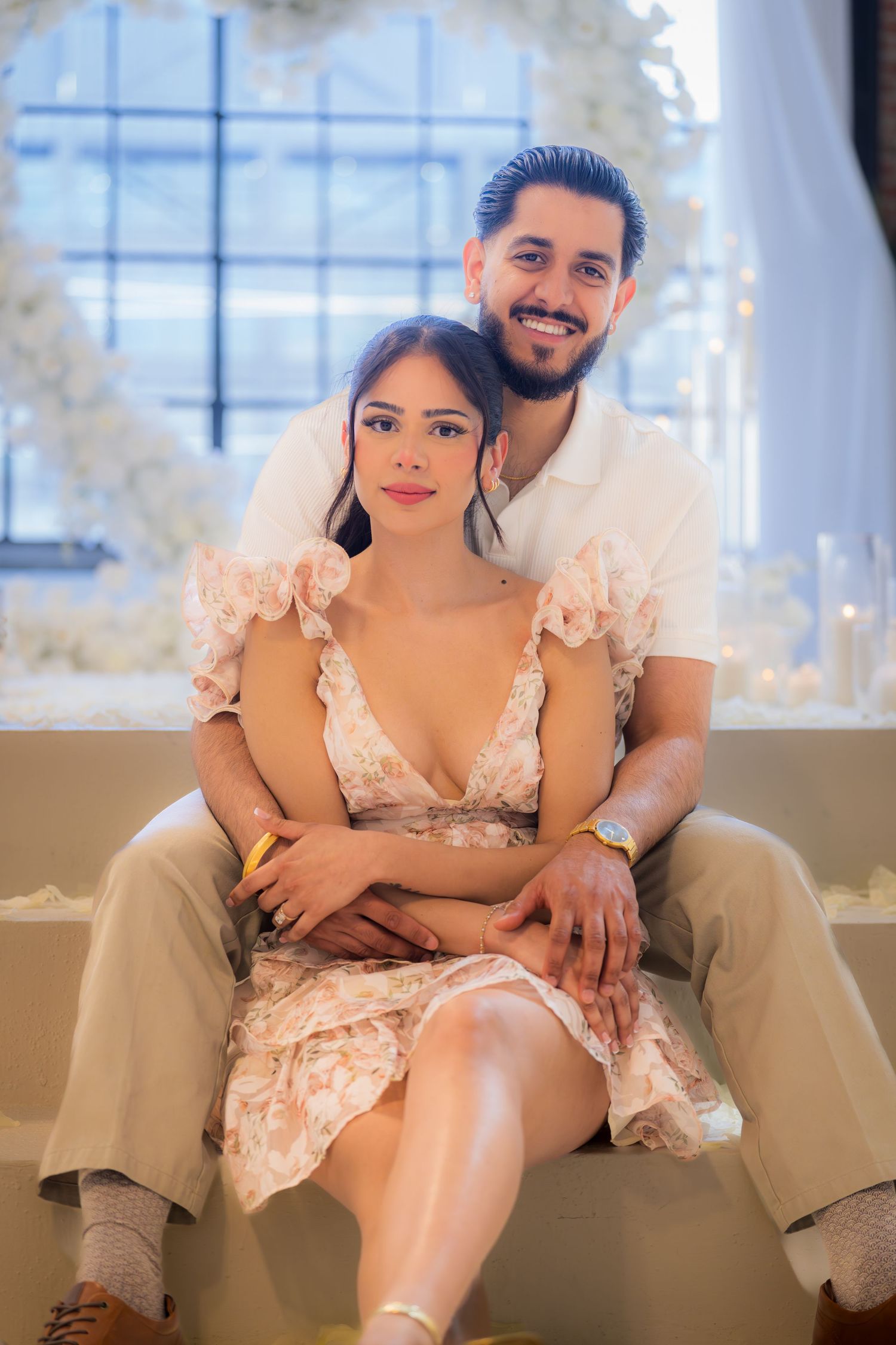 Engaged couple posing together in front of elegant white floral arrangement backdrop.