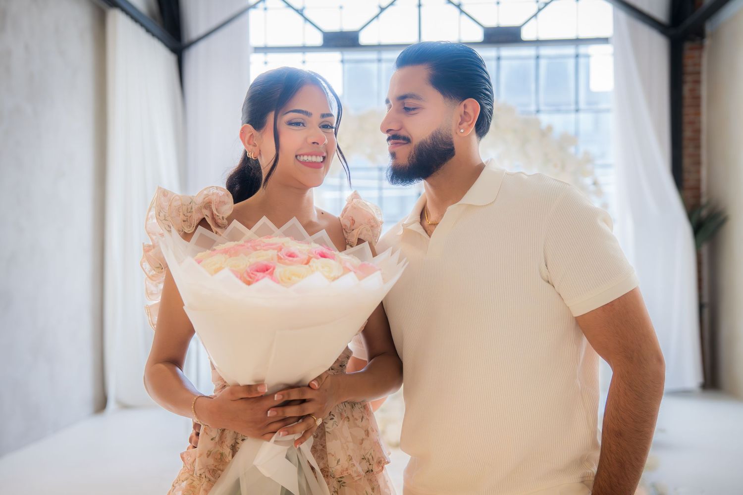 Smiling couple sharing a large bouquet of pink and white flowers indoors.