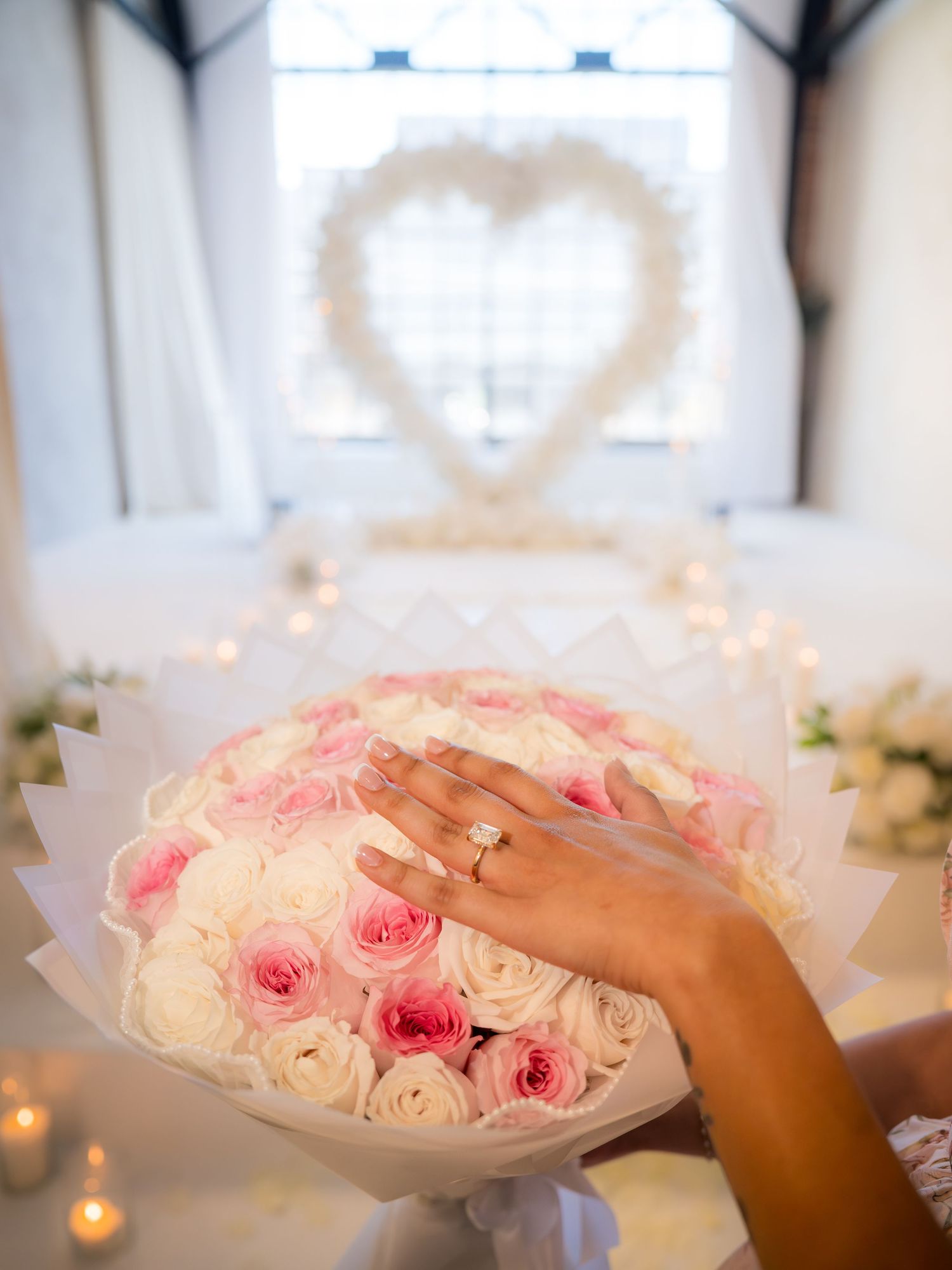 Hand with diamond engagement ring resting on pink and white rose bouquet with heart decor backdrop.