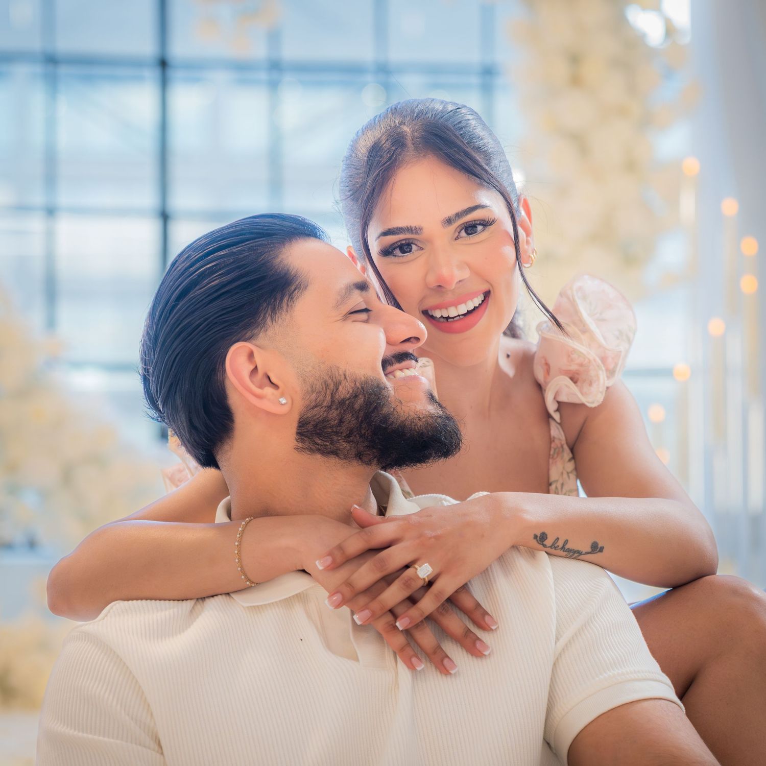 Joyful engaged couple embracing and smiling together in front of white floral heart arch backdrop.
