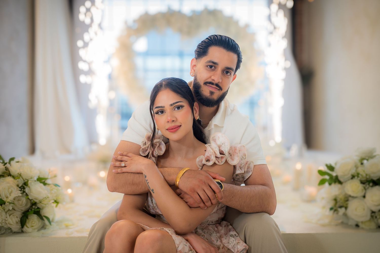 Couple cuddling in front of illuminated heart arch and white floral wedding decor.