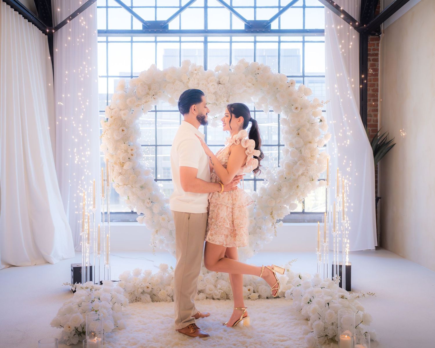 Couple sharing a tender moment beneath a white floral heart arch with twinkling fairy lights.
