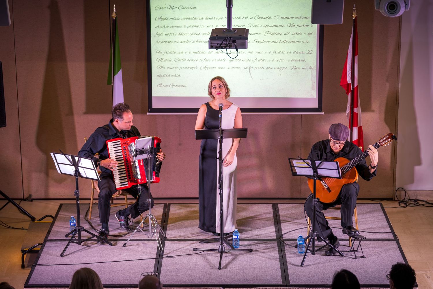 A woman in white performs on stage with an accordion player and guitarist, flanked by Italian and Canadian flags.