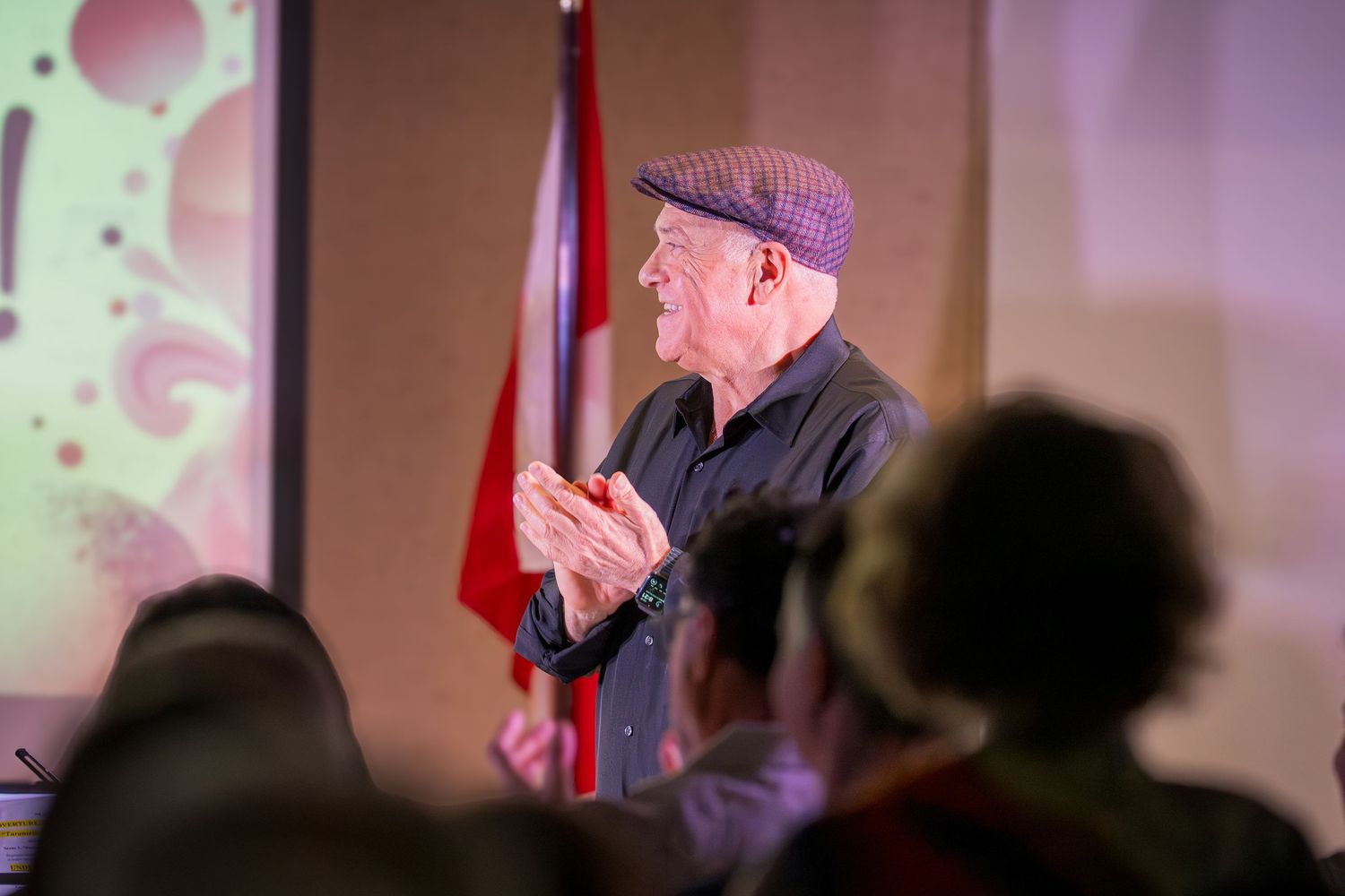 An elderly man in a flat cap applauds in front of a Canadian flag at an indoor cultural event.