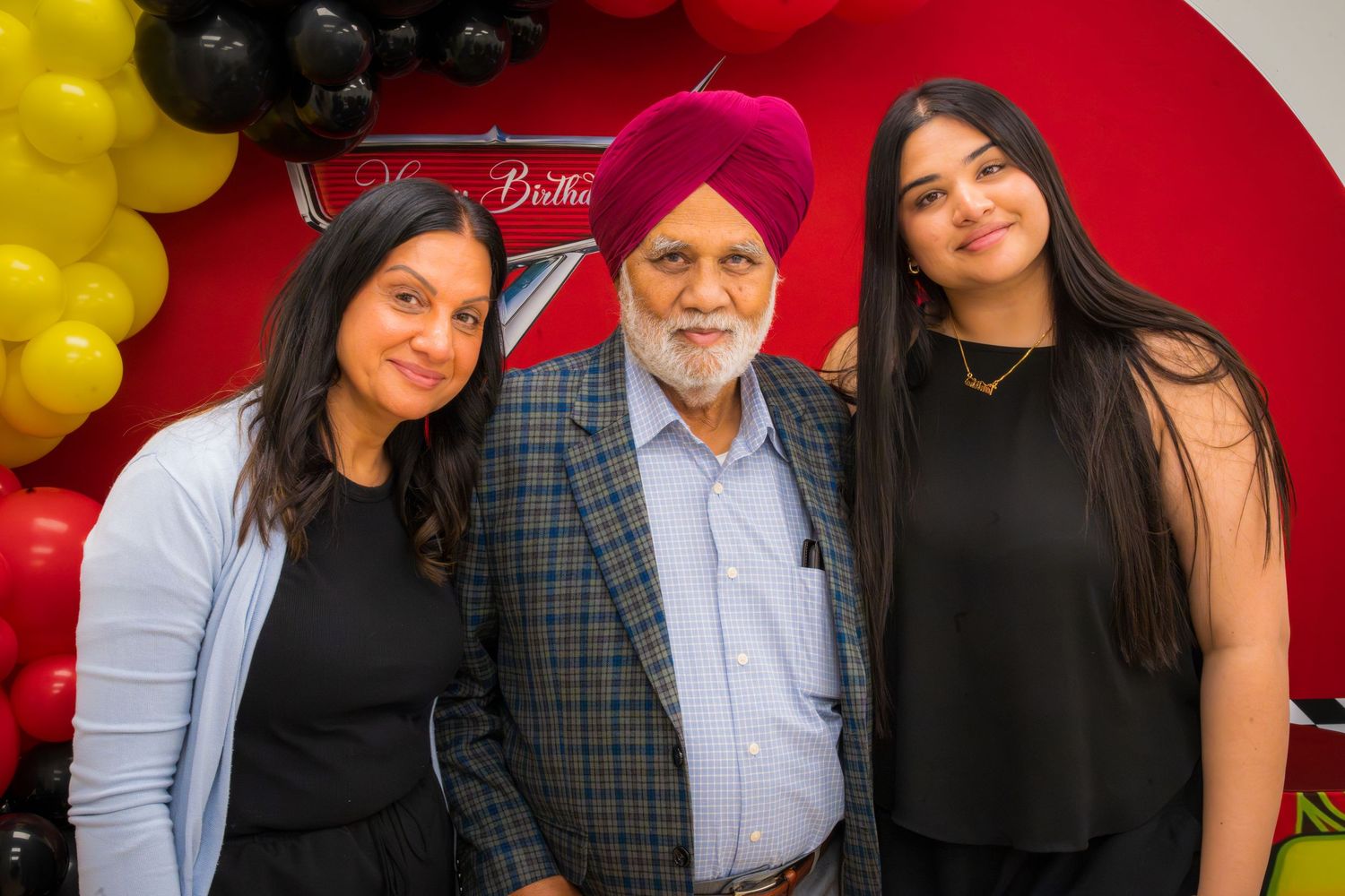 Three people smiling at a birthday celebration with colorful balloons in the background.