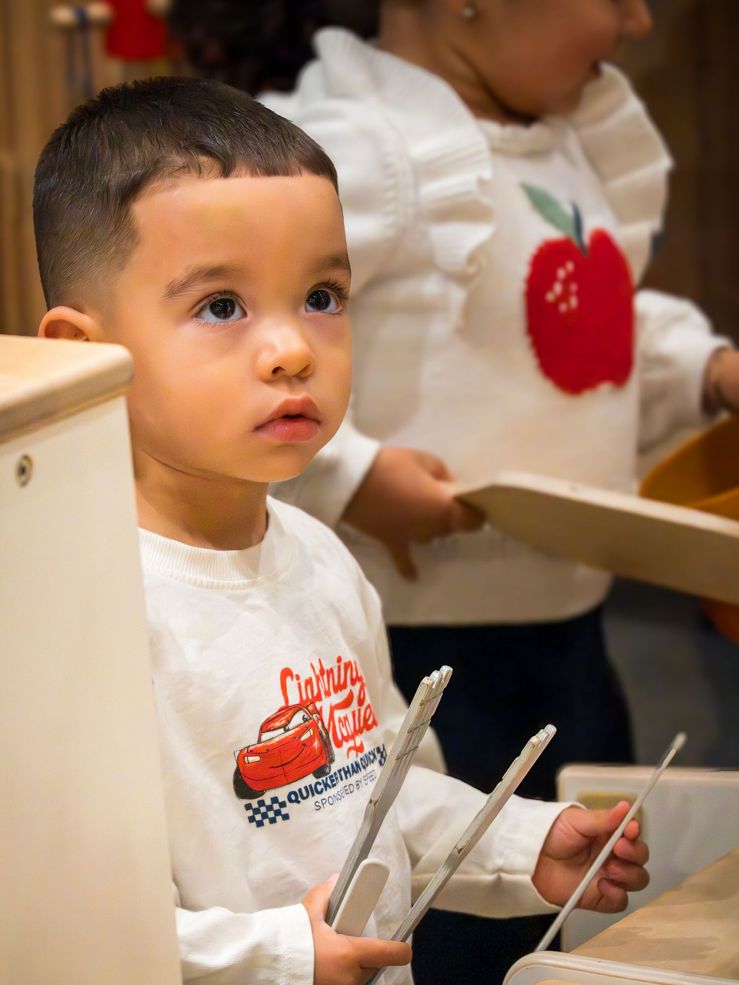 A young boy in a Lightning McQueen shirt holds drumsticks while standing near wooden furniture.
