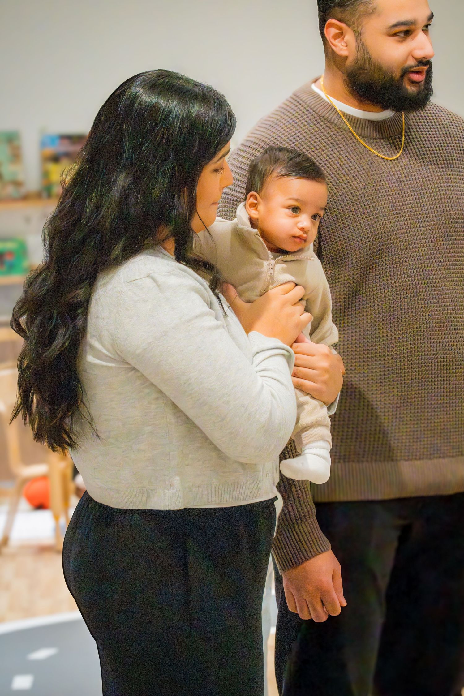 Woman in gray sweater holding a baby while a bearded man stands beside her indoors.