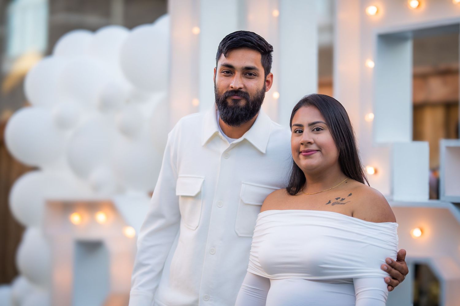 Couple in white outfits posing together in front of white balloon decor and illuminated marquee letters at baby shower.