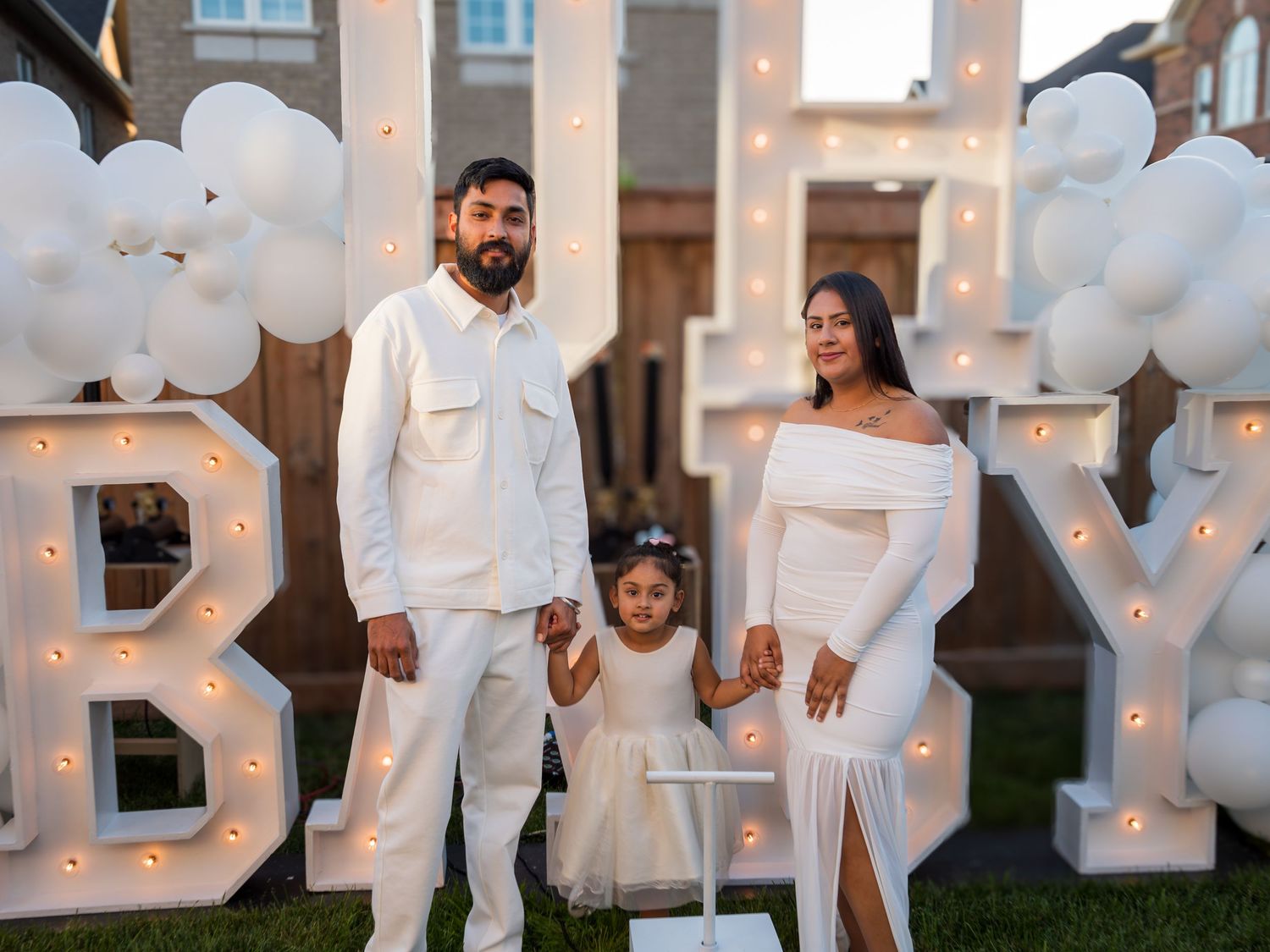 Family of three in white outfits stands before illuminated BABY marquee letters with white balloons.