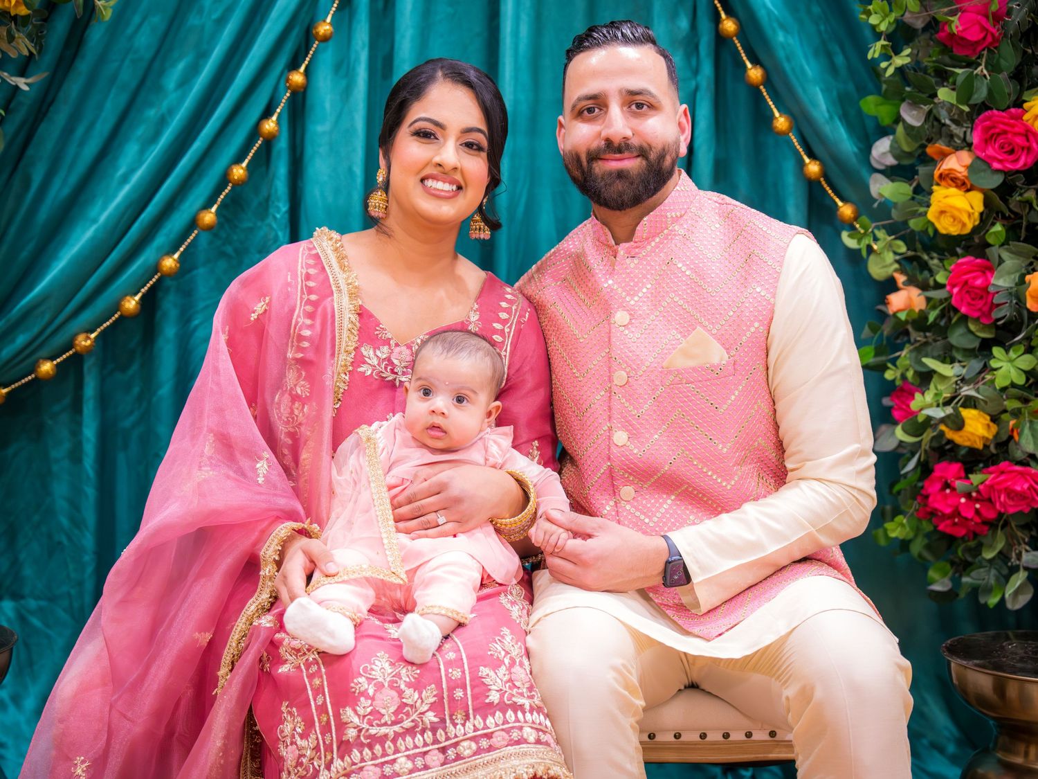 Indian family in matching pink outfits posing with newborn baby at a colorful floral celebration.