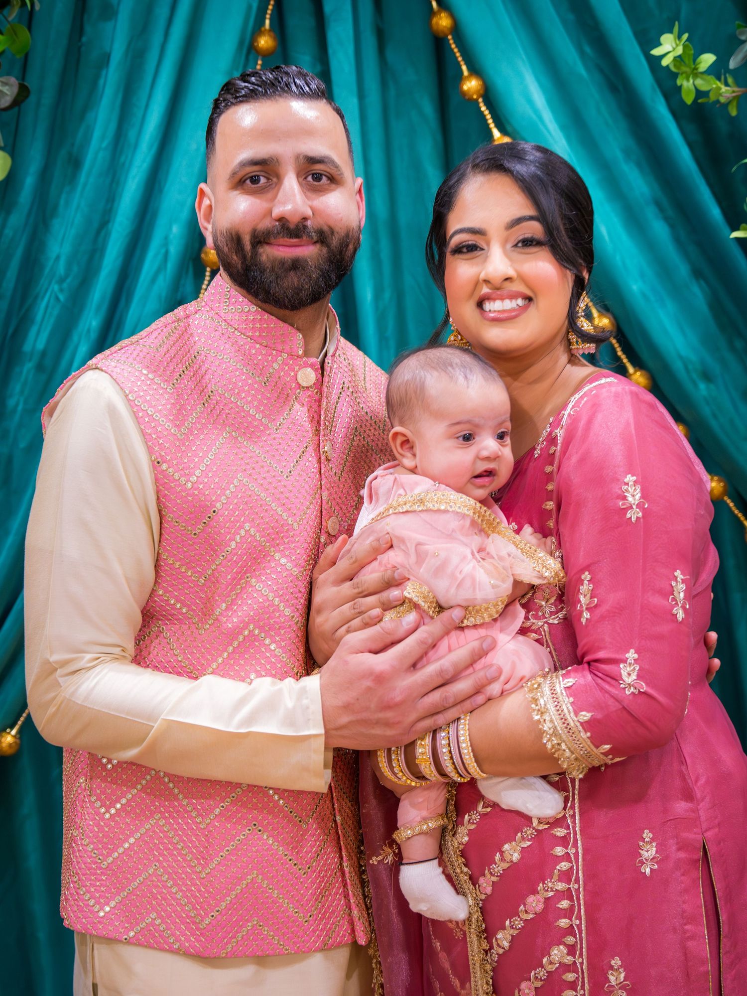 Couple in matching pink Indian traditional outfits holding their baby at a South Asian celebration ceremony.