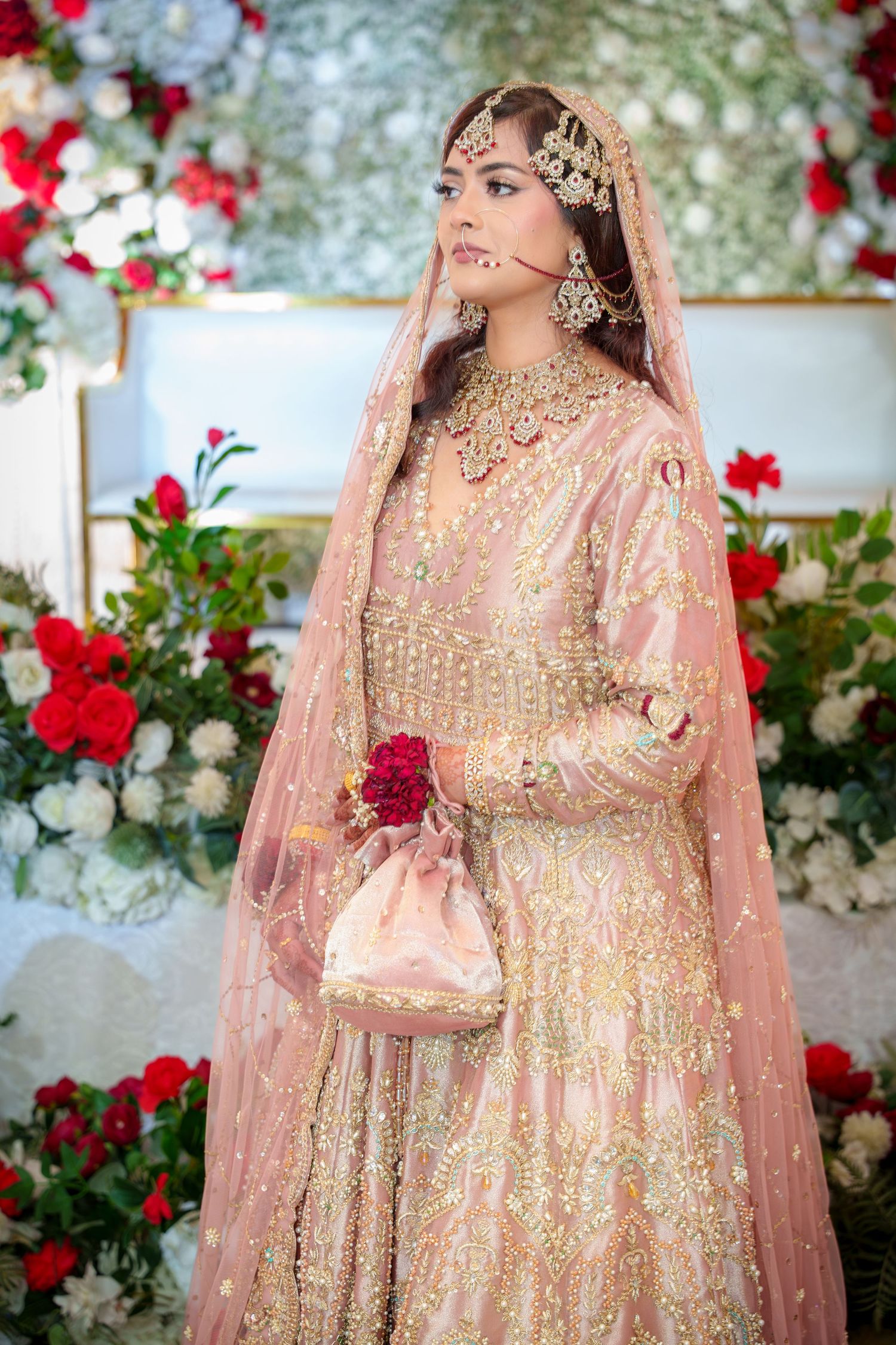 South Asian bride in pink and gold embroidered lehenga poses against backdrop of red and white flowers.