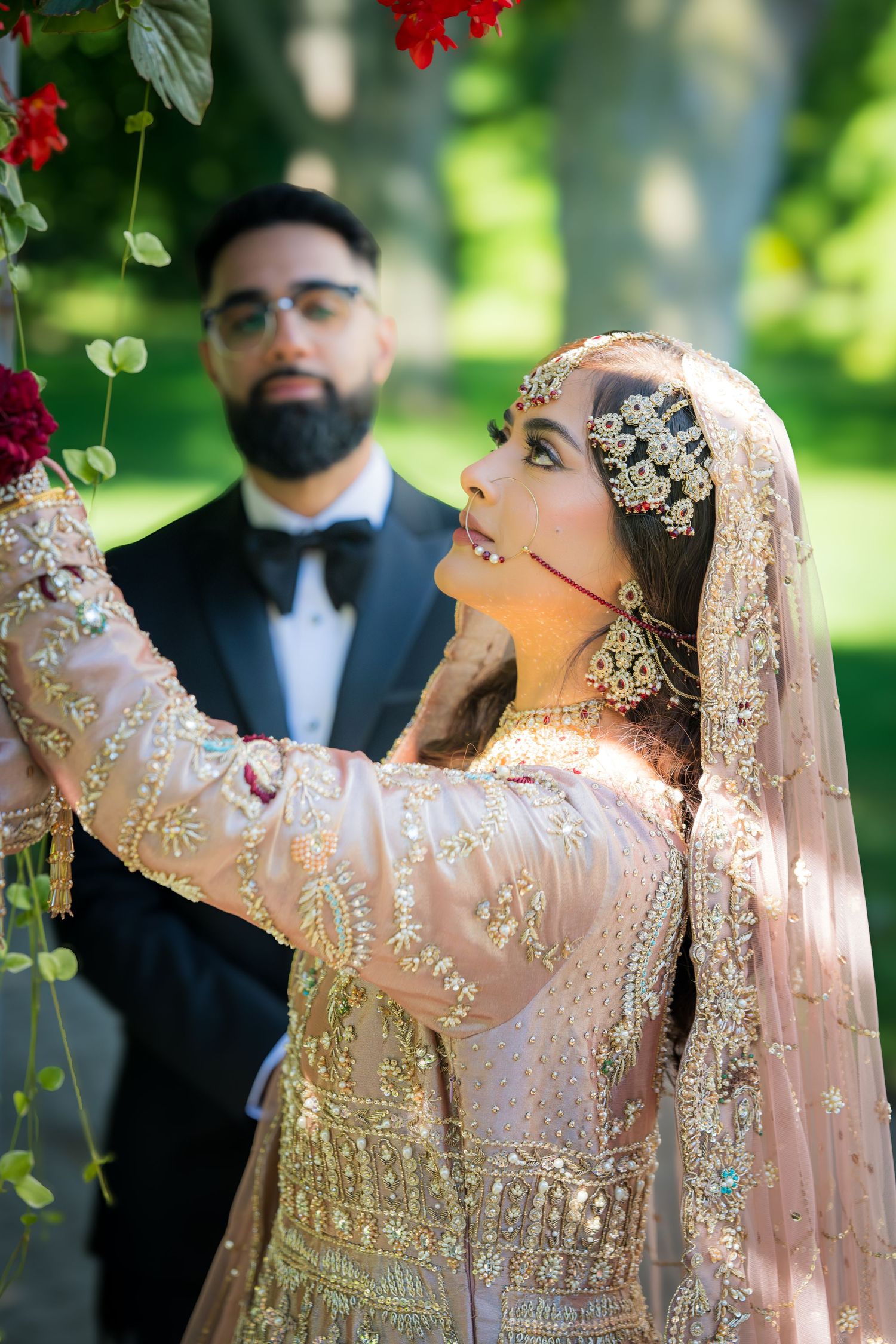 Traditional South Asian wedding couple poses with red flowers as bride reaches up in ornate pink and gold lehenga.