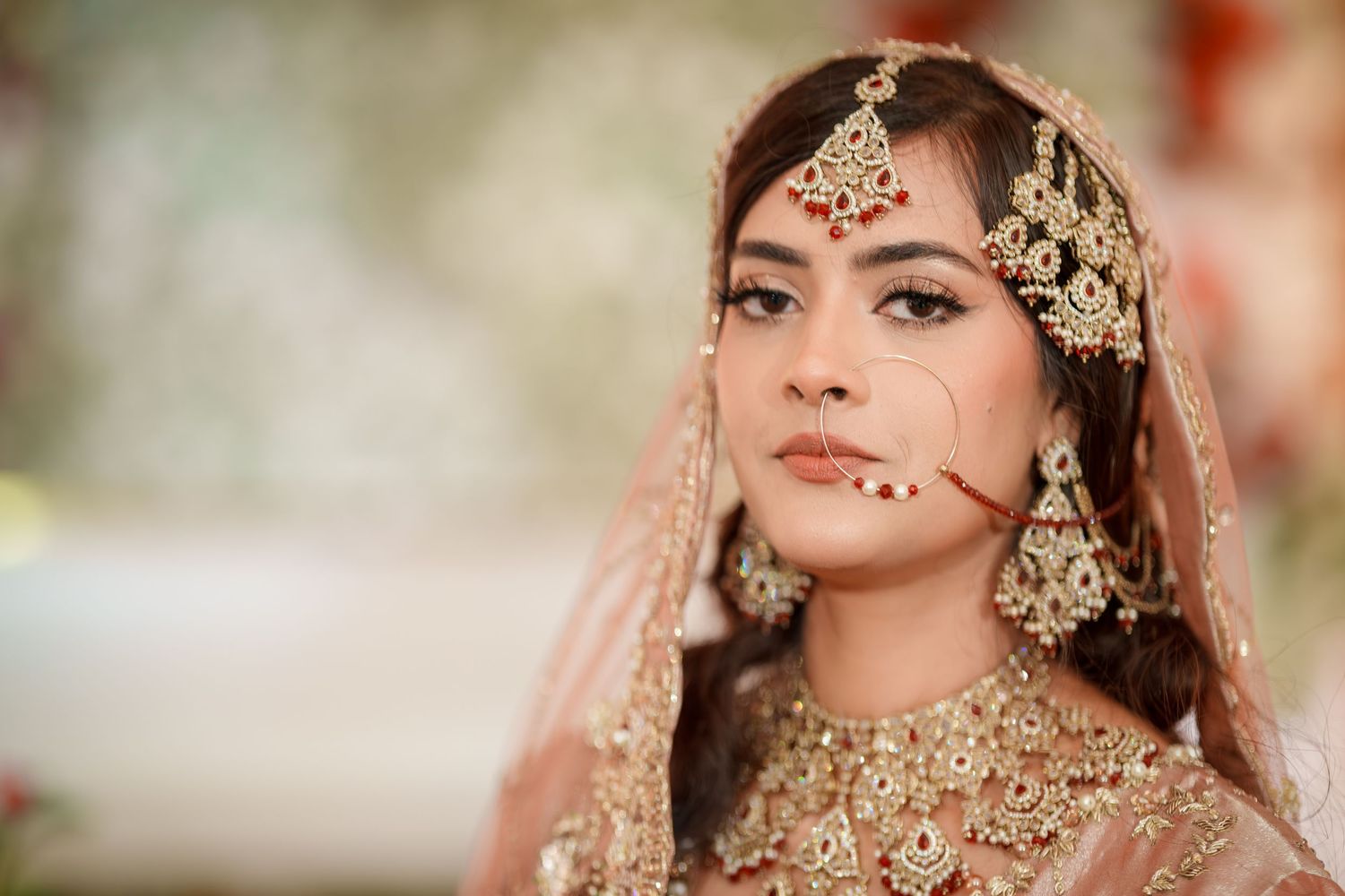 Bride wearing ornate gold jewelry and traditional South Asian bridal makeup in soft lighting.