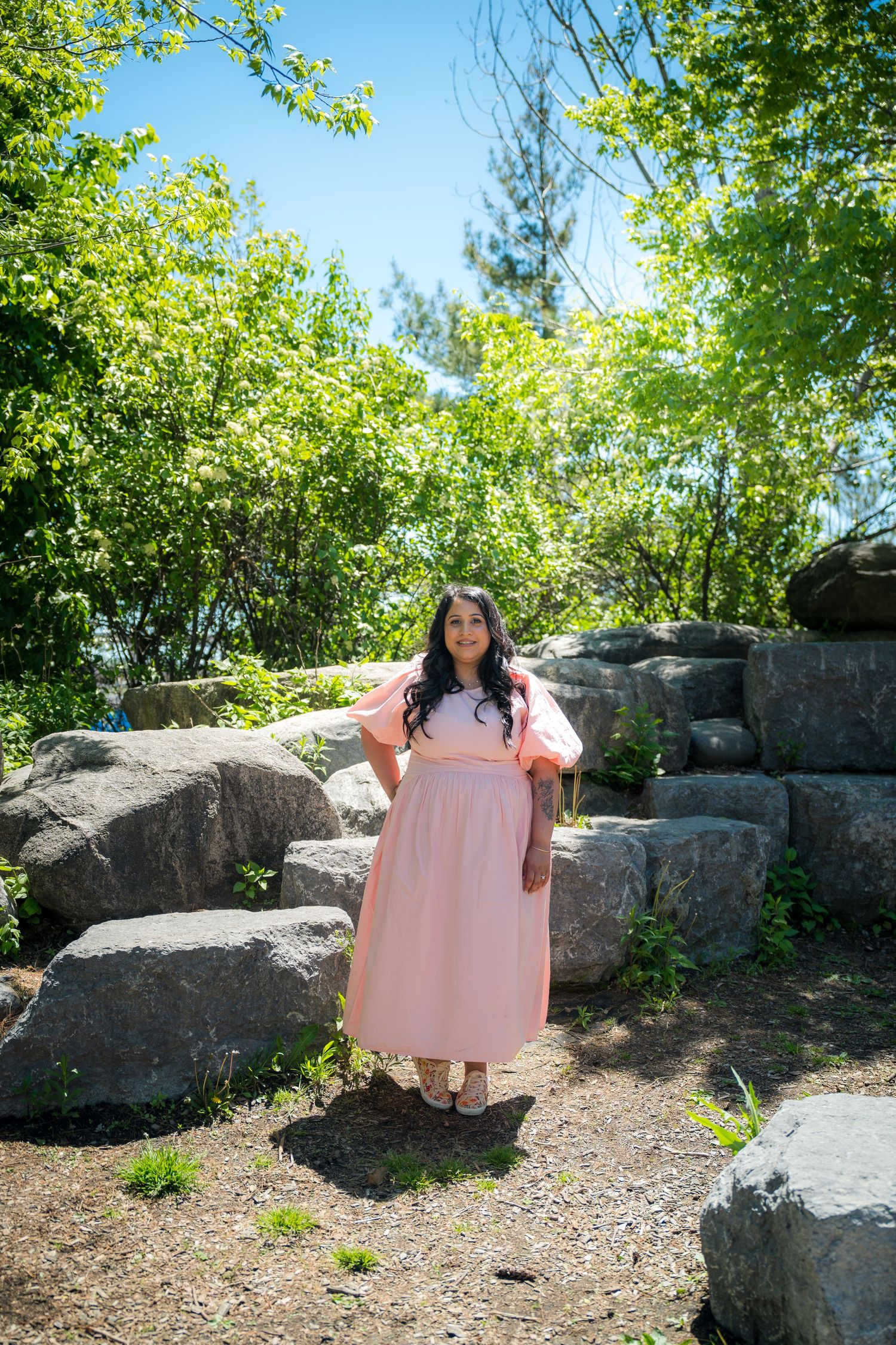 Woman in pink maxi dress standing among rocks and green trees in a sunny park.