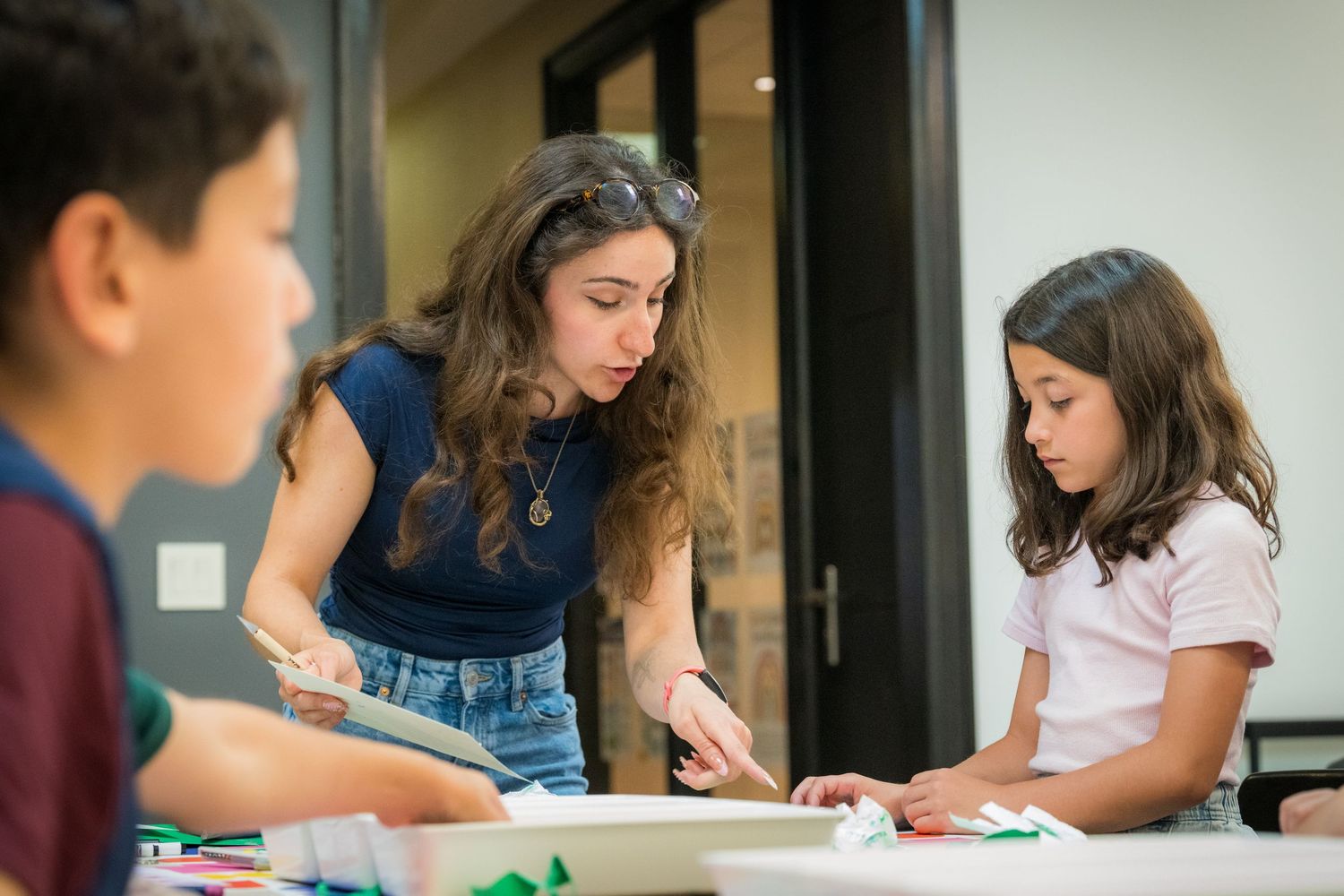 Female teacher leans over to help young girl with art project at a colorful craft table.
