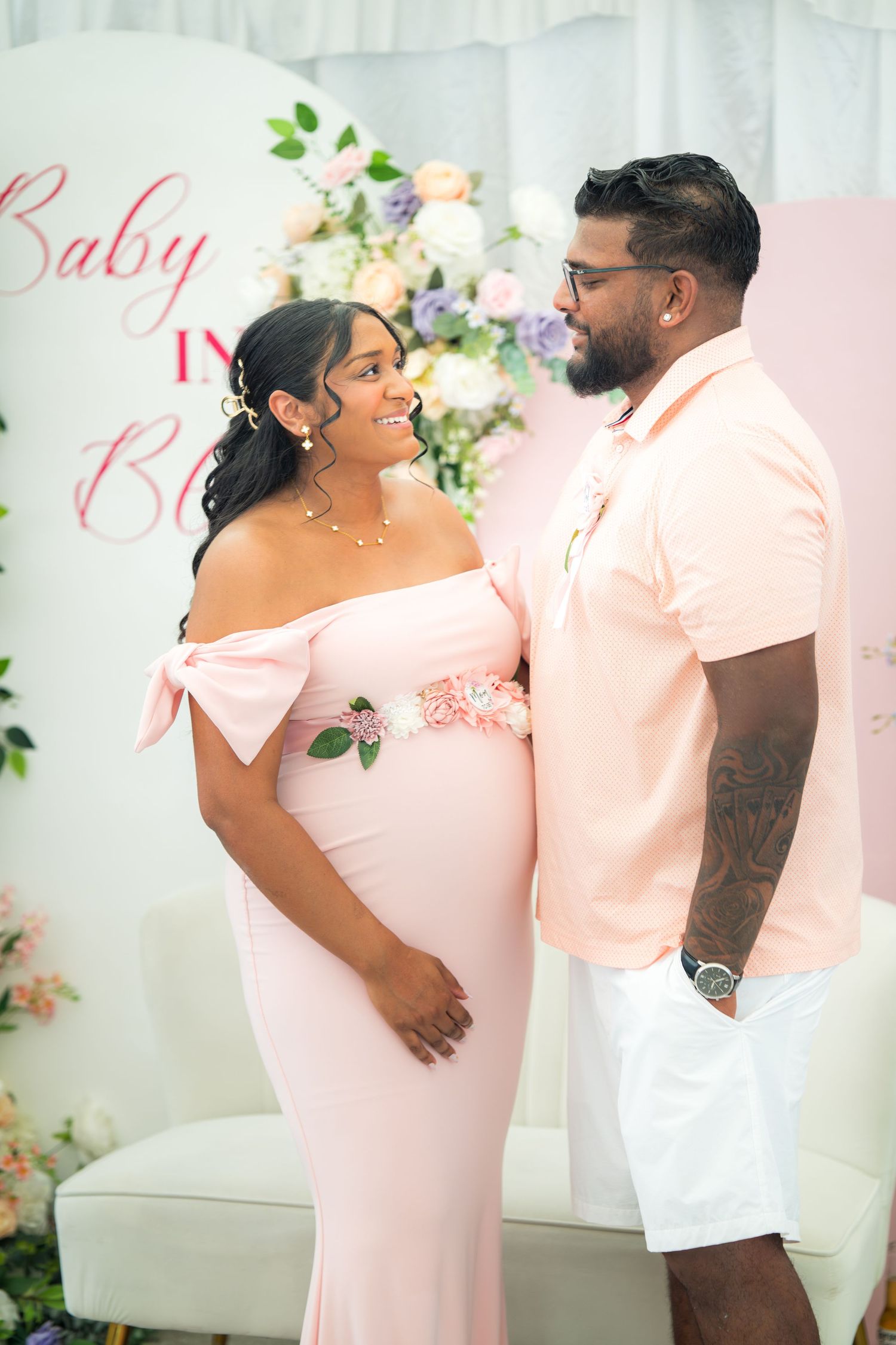 Expectant couple shares loving glance at baby shower with floral wall decor and pink theme.