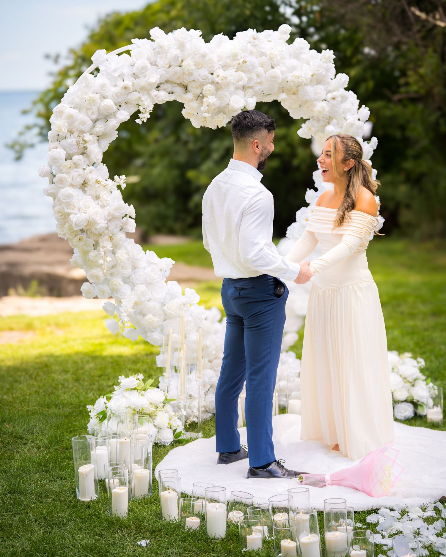 A romantic outdoor wedding ceremony setup with a white floral arch and glowing candles lining the aisle.