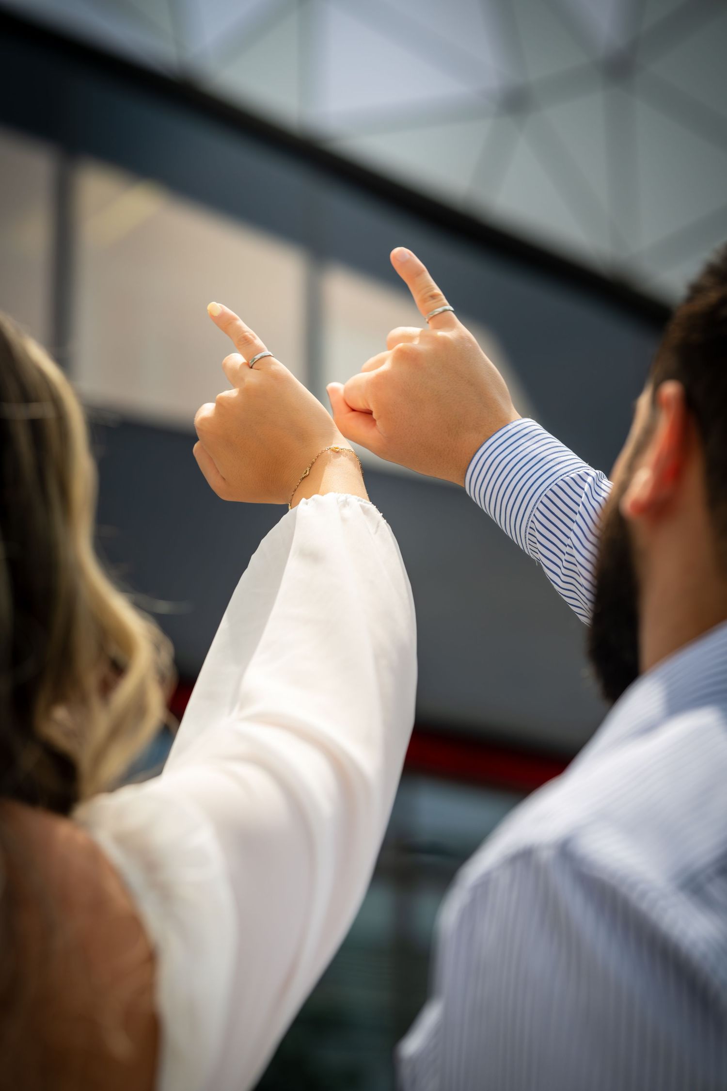 Two people point upward while posing against an architectural background.