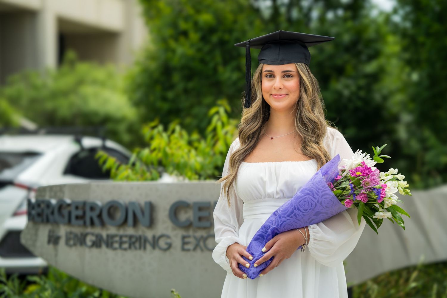 Graduate poses with diploma and flowers outside university engineering building.